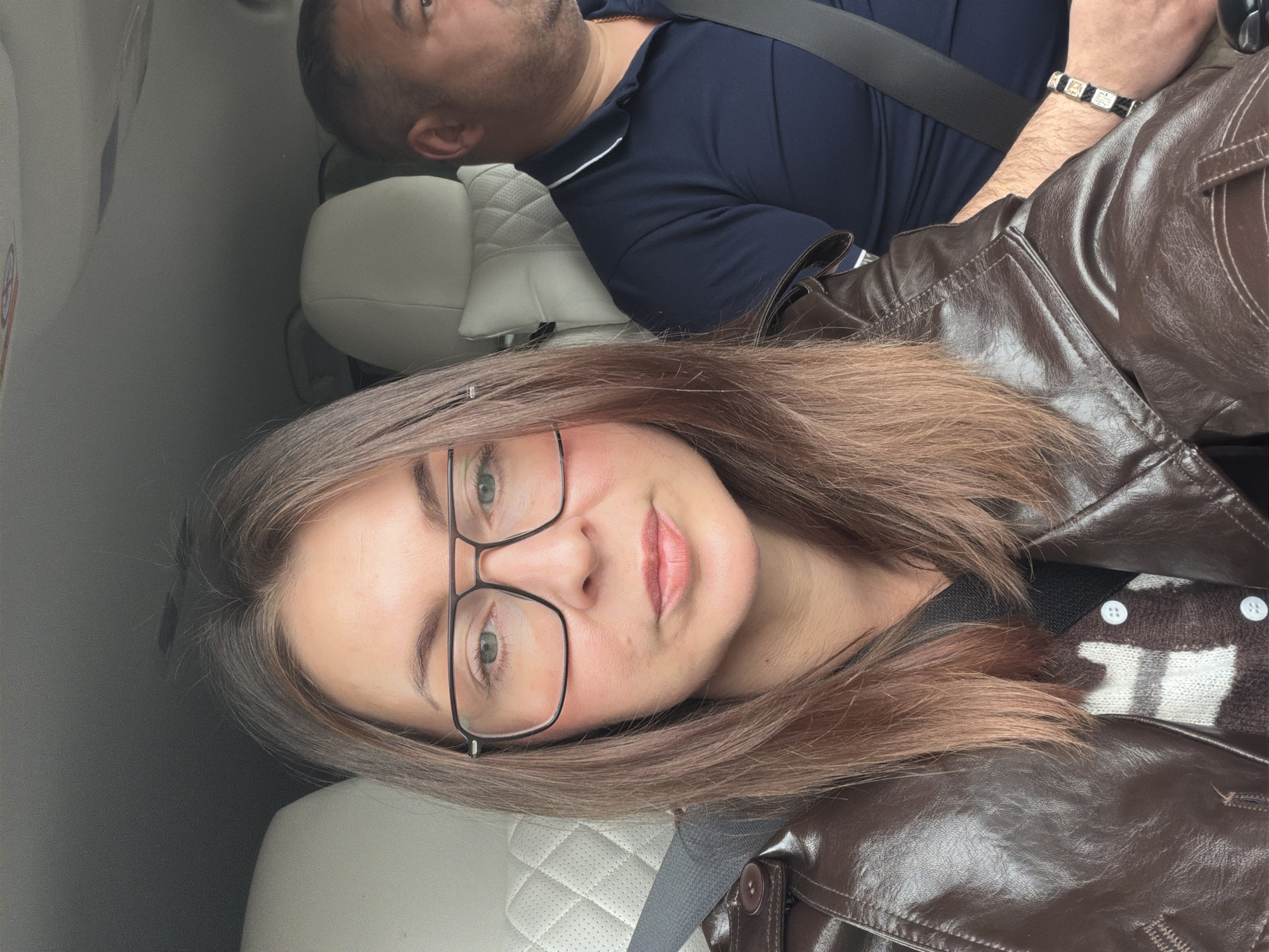A woman is taking a selfie in the back seat of a car. She has fair skin, brown hair, and is wearing glasses. She is wearing a brown leather jacket. In the background, a man is sleeping or resting in the seat next to her. He is wearing a dark blue collared shirt. The car seats are light colored. The environment suggests travel; however, the photo provides no information related to the city of Поварёнки, Russia.