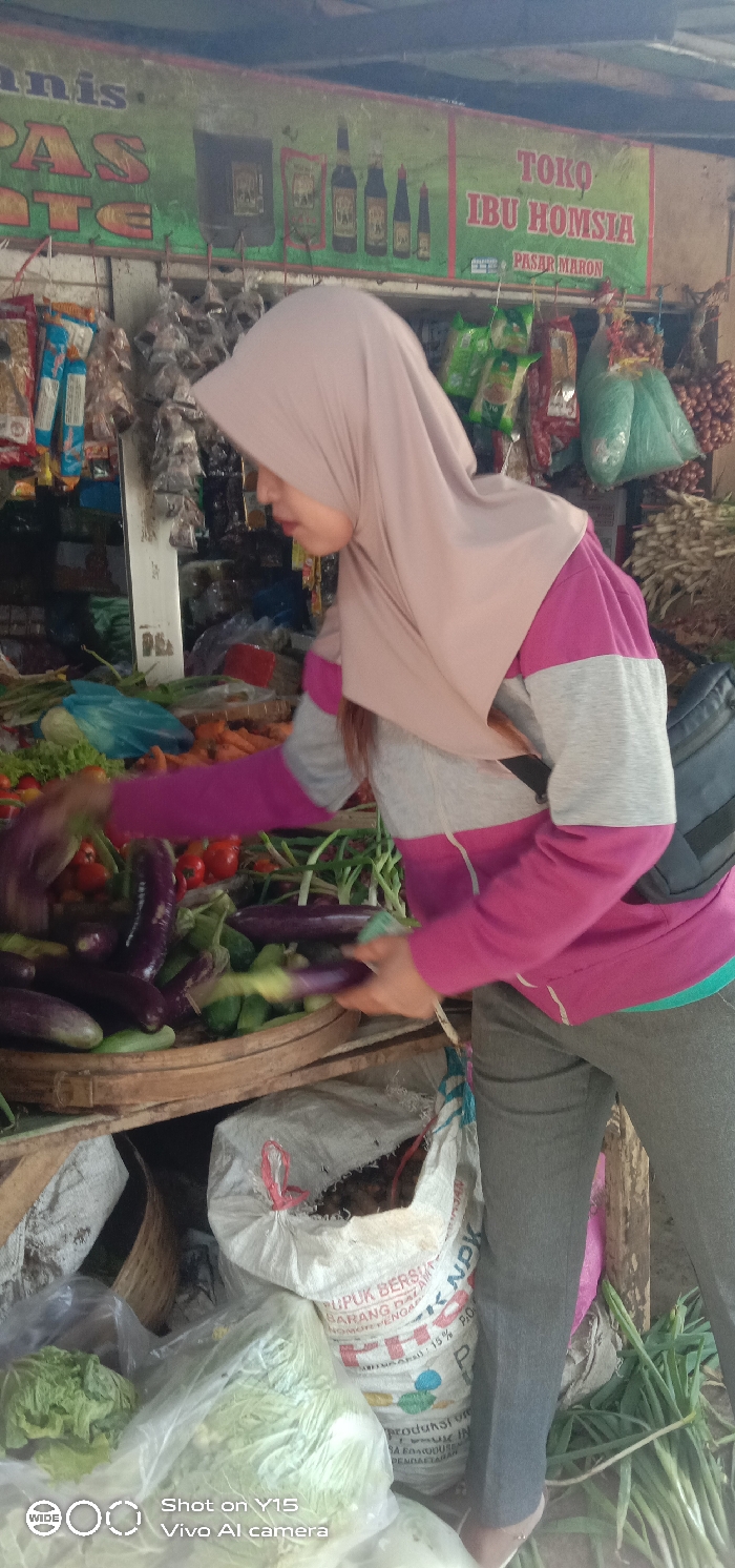 The image depicts a retail market stall within Pasar Maron (Maron Market) in Maron, Indonesia. A female individual, positioned in a three-quarter profile, faces left and interacts with a vegetable display. She wears a light pink hijab, a long-sleeved shirt featuring prominent magenta and light gray horizontal stripes, dark gray trousers, and light-colored shoes. A black sling bag is worn across her back. Her hands are engaged within a large, round wooden basket containing numerous dark purple eggplants and green cucumbers, indicating selection or arrangement of produce.
In the background, a green banner above displays "TOKO IBU HOMSIA PASAR MARON" along with illustrations of various bottled condiments. A partial banner "nis PAS ATE" is visible to the left. Hanging from hooks are numerous packaged items in clear plastic bags, possibly snacks or spices, and larger bundles of produce such as red onions. Behind the vegetable basket, piles of red tomatoes and orange carrots are visible. Below the primary vegetable display, several large white woven polypropylene sacks are present, one partially labeled "PUPUK BERSAHABAT" and "NPK," containing other market goods. Clear plastic bags containing light green leafy vegetables, possibly cabbage or lettuce, are also on display at ground level.