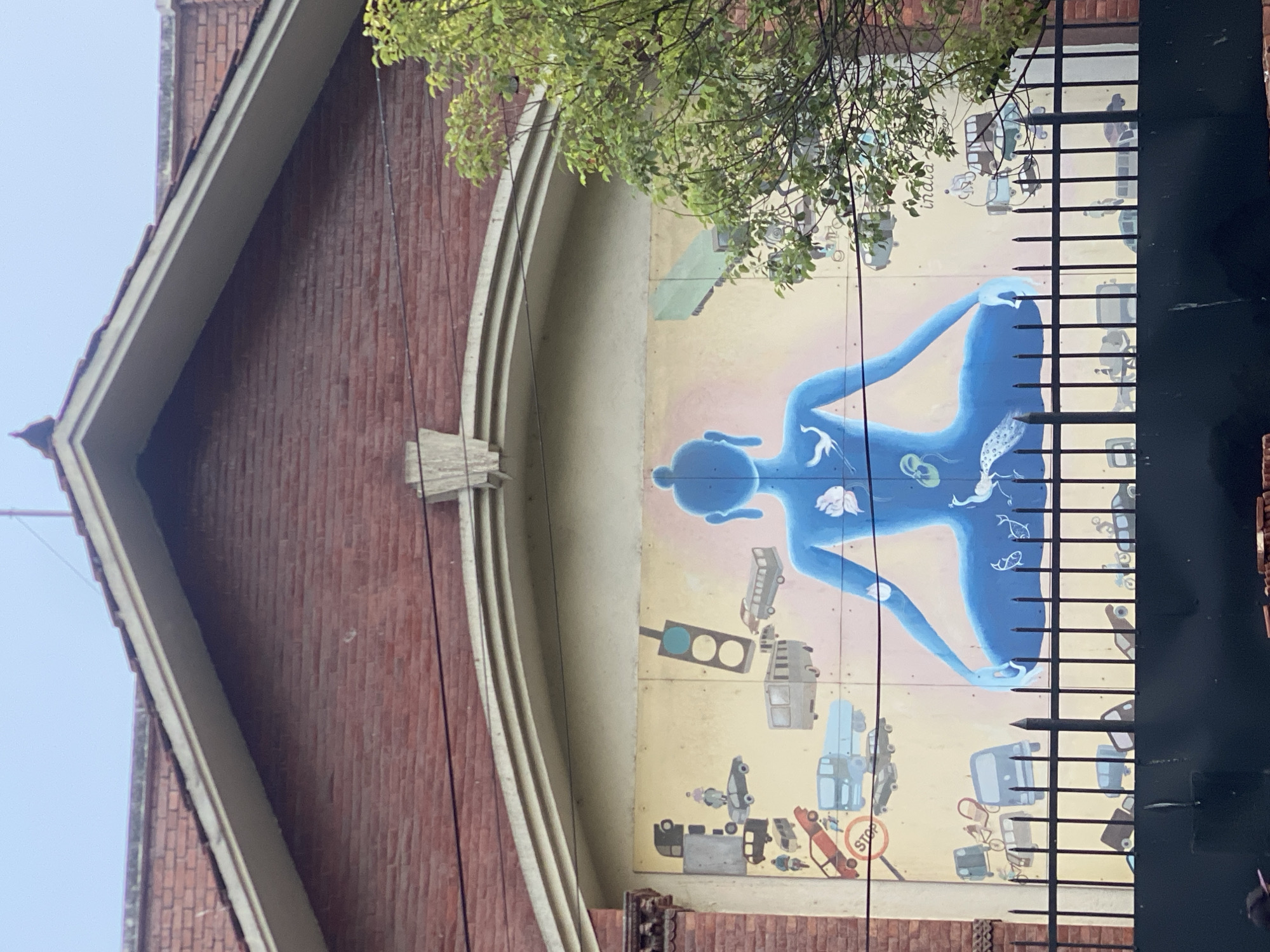A mural is painted on the side of a brick building in Kathmandu Metropolitan City, Nepal. The mural depicts a large, blue, silhouetted figure in a meditative pose, with various forms of transportation, including cars, buses, and motorcycles, depicted around and within the figure. Birds and a flower are also integrated into the design. A black metal fence runs in front of the mural, and a green tree partially obscures the right side of the artwork. The building has a red brick facade and a sloped tiled roof. The sky is overcast.