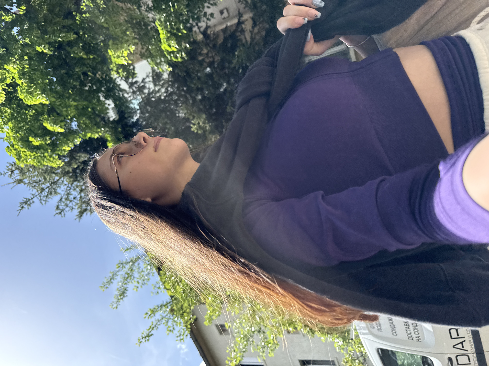 The image shows a woman in sunglasses and a purple shirt, standing under trees in Sofia, Bulgaria. She's holding a black hoodie and looking up towards the sky. The background includes a building, a clear blue sky with some clouds, and a delivery van with "DAP" written on it. The text on the van is in Bulgarian, reading "ДОСТАВКИ НА СОФИЯ". The location appears to be outdoors on a sunny day.