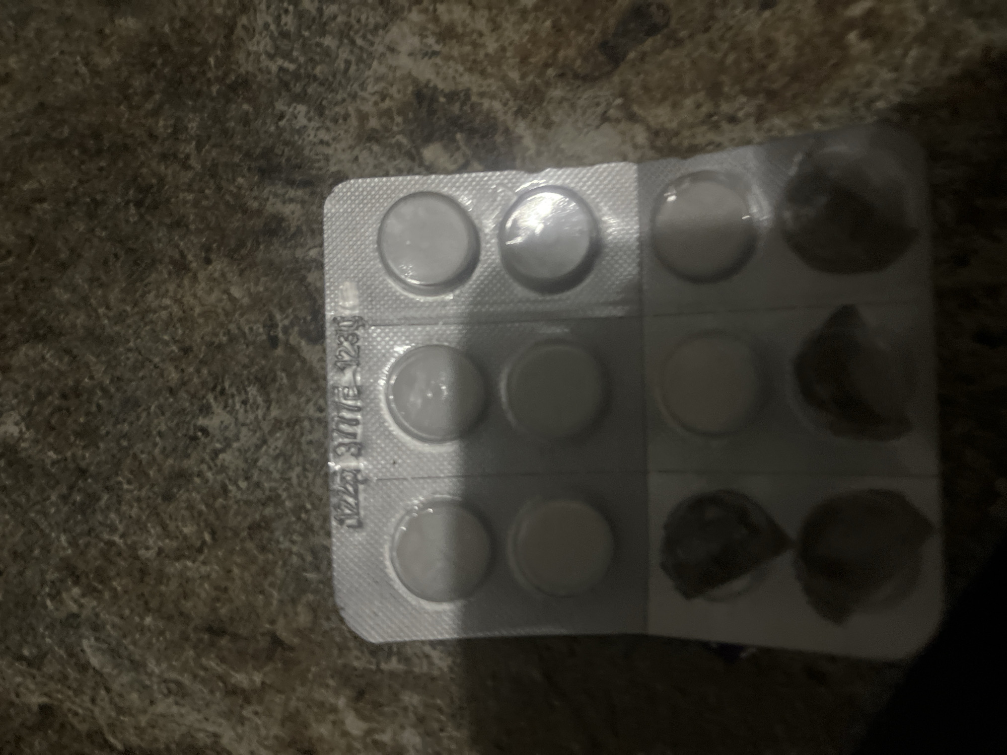 A blister pack of pills, with some pills removed, is lying on a textured, brown surface, possibly a carpet or blanket. The blister pack is silver and contains nine circular depressions, six of which hold white pills. The remaining three depressions are empty, with dark residue visible in two of them. Embossed on the foil of the blister pack are the numbers "1223 3/77E 1230". The image is taken from a slightly elevated angle, with the background blurred and dark. The location in Jalingo, Nigeria is not discernible from the image.