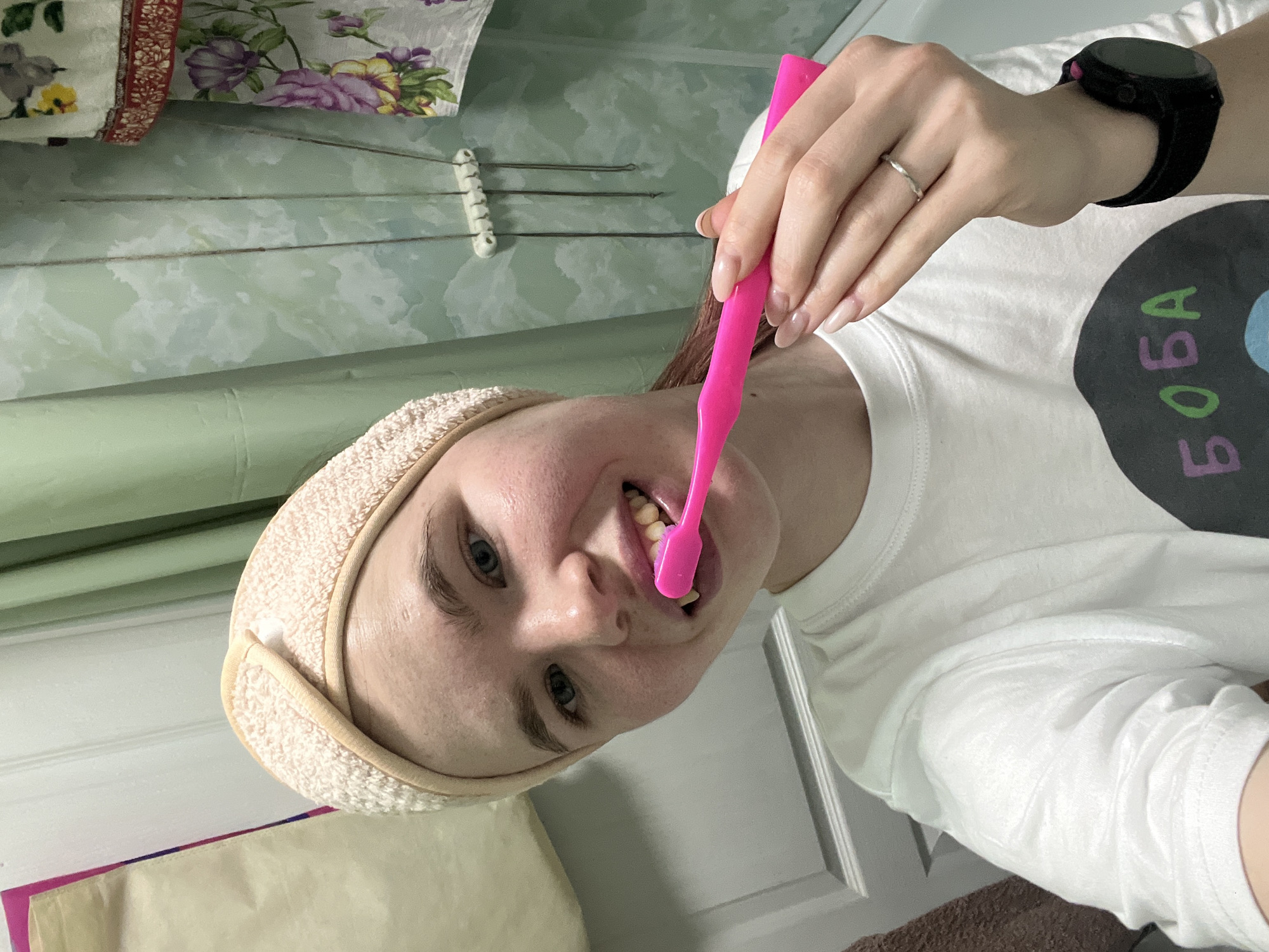 A woman with fair skin and light brown hair, wearing a beige headband and a white t-shirt with the word "БOБA" printed on it, is brushing her teeth with a pink toothbrush. She has a black smartwatch on her left wrist and a ring on her finger. The background shows a light green wall with floral patterns, a row of metal hooks, and a partially visible door. The location of the photo may be in Chaikovsky, Russia.