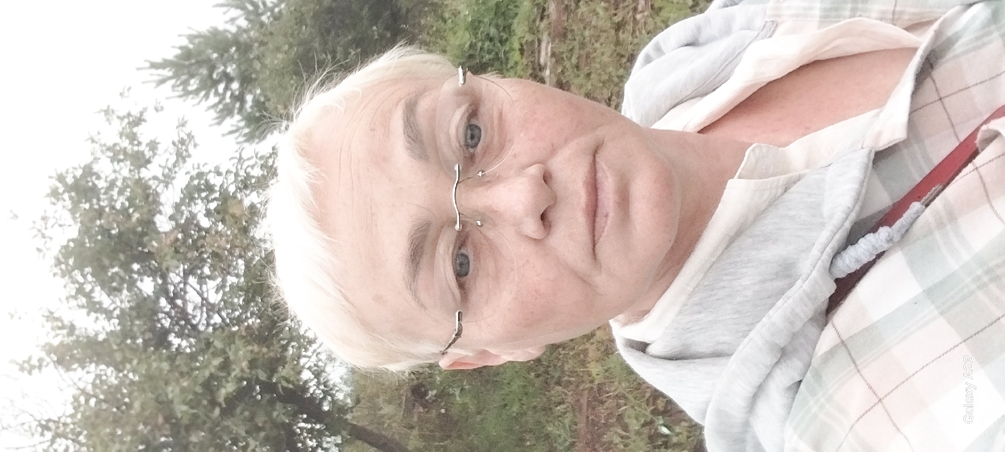 The image is a headshot of a fair-skinned woman with short, blonde hair and blue eyes, wearing rimless glasses. She has freckles on her face. She is wearing a light-colored collared shirt under a light gray hooded jacket. The background consists of greenery, with trees and grass, suggesting an outdoor setting, possibly in a rural area. The lighting is overcast. A watermark at the bottom left of the image reads "Galaxy A23". The location is specified as Novotroitskoe, Russia, though no specific city landmarks are visible.