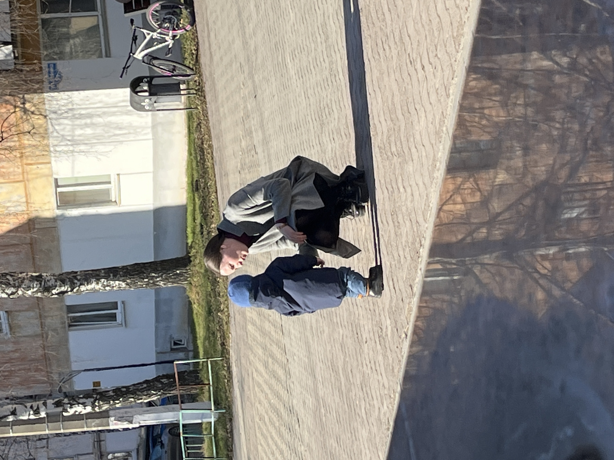 The image captures an outdoor scene in Chaikovsky, Russia, featuring a woman and a young child on a paved surface under bright sunlight. The main image is rotated 90 degrees clockwise; when viewed upright, the scene depicts a sidewalk or courtyard alongside residential buildings.
In the foreground, a woman, dressed in a light grey coat over dark clothing and a dark red turtleneck, is bending down to eye level with a child. Her dark hair is styled in a bob. The child, wearing a navy blue jacket, blue jeans, and a blue knit cap, stands facing the woman, suggesting an interaction or conversation between them.
The ground is a light-colored paved surface with distinct horizontal lines, indicating individual pavers. Long, sharp shadows are cast across the pavement and a nearby wall, characteristic of early morning or late afternoon sun. To the left of the figures, a white bicycle with pink details on its wheels and seat is parked next to a black metal street bench or trash receptacle.
In the background, there are multi-story residential buildings. One building is painted a light grey or white, with visible windows. Another building to its left has a brownish, possibly stuccoed, facade. Several trees with bare branches stand between the paved area and the buildings, some appearing to be birch trees. A patch of green grass is visible to the right. No discernible text is visible on the buildings or objects. The overall atmosphere appears calm and bright.