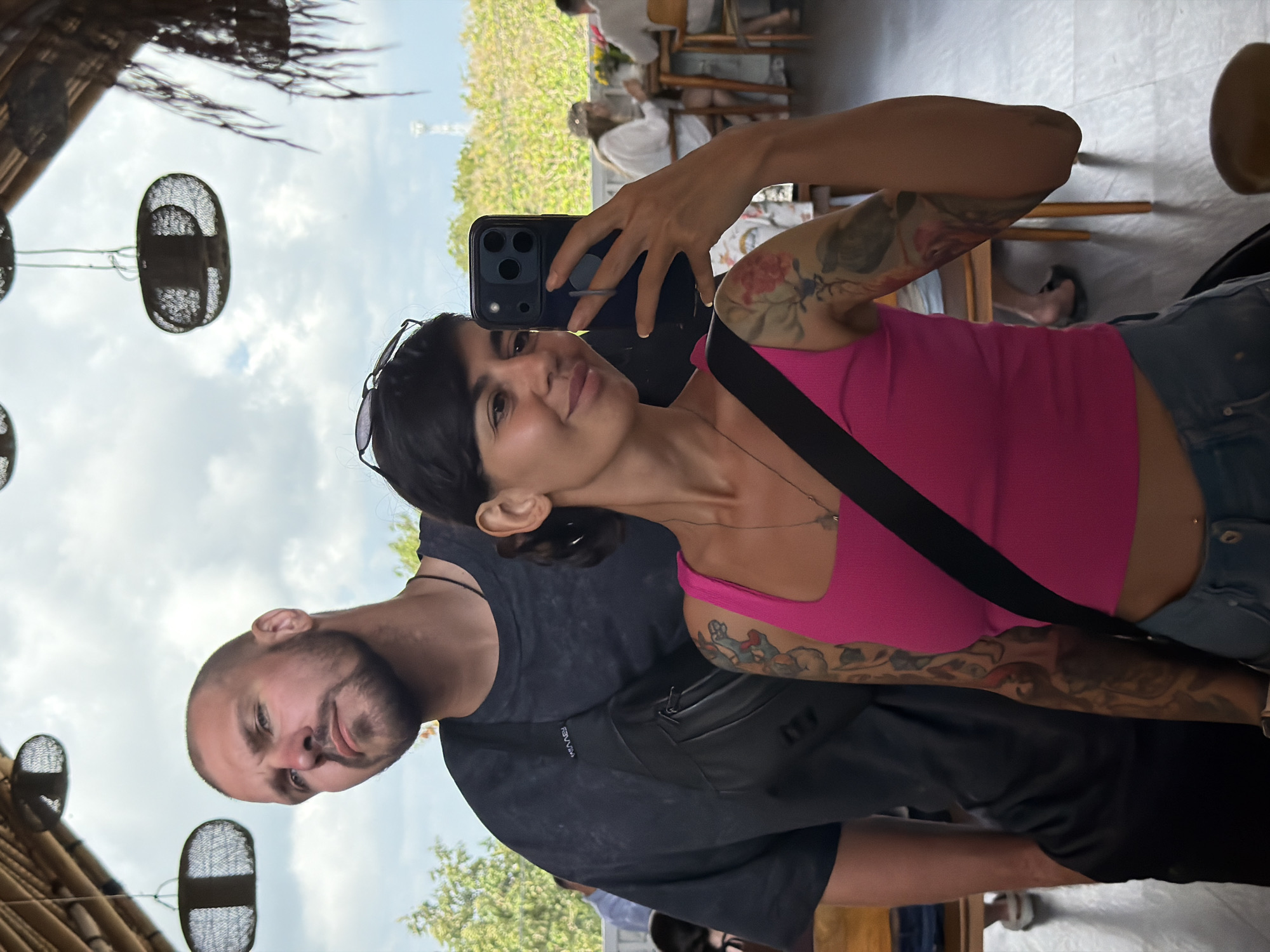 In Kuta Selatan, Indonesia, a couple takes a selfie in a restaurant with a thatched roof and hanging woven lamps. The man, in a dark grey t-shirt with a fanny pack across his chest, stands behind the woman. He has a shaved head and a beard, and he is looking to the left of the camera. The woman, in a bright pink tank top and denim shorts, holds a smartphone to take the picture. She has dark hair pulled back and is smiling at the camera. Her left arm, visible in the frame, is adorned with floral tattoos. In the background, other patrons are seated at tables, and lush greenery is visible through an open window.