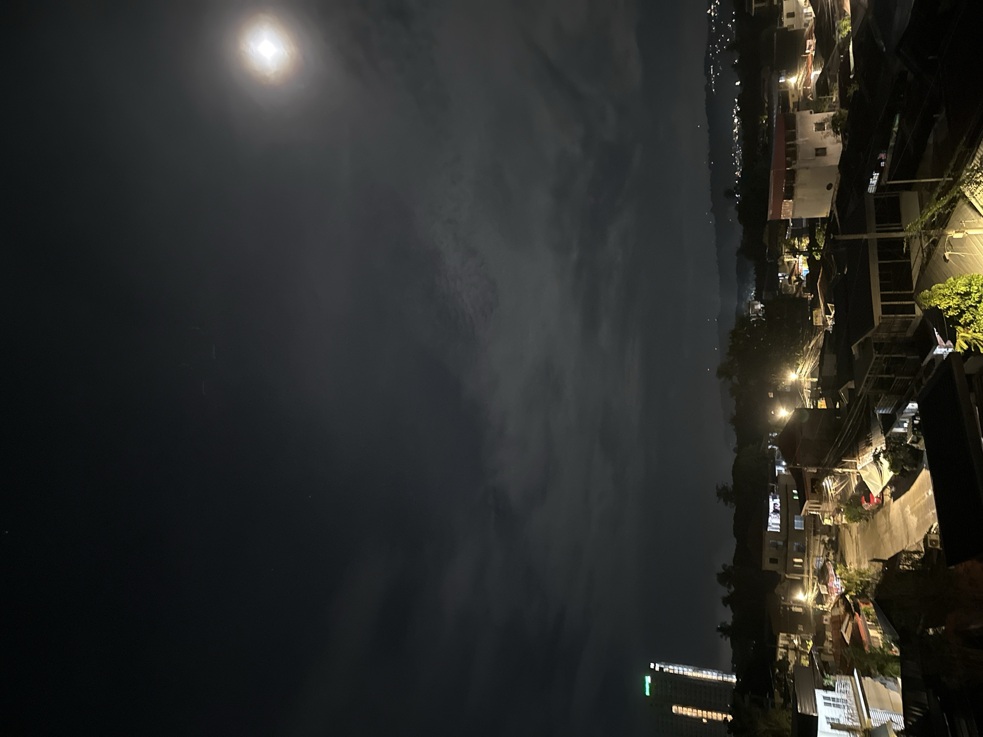 This photograph captures an aerial view of a residential area in Davao City, Philippines, at night. The sky is dark with scattered clouds, and a bright, somewhat hazy moon is visible in the upper right quadrant. A tall, illuminated skyscraper stands out in the background on the left, displaying green signage that reads "ONE OASIS." Below the sky, a dense cluster of houses and buildings with illuminated windows fills the frame. Streets are visible, some with streetlights, and a few vehicles, including a red car, are parked. Trees and foliage are interspersed among the buildings. The overall impression is of a moderately populated urban landscape under a moonlit night sky.