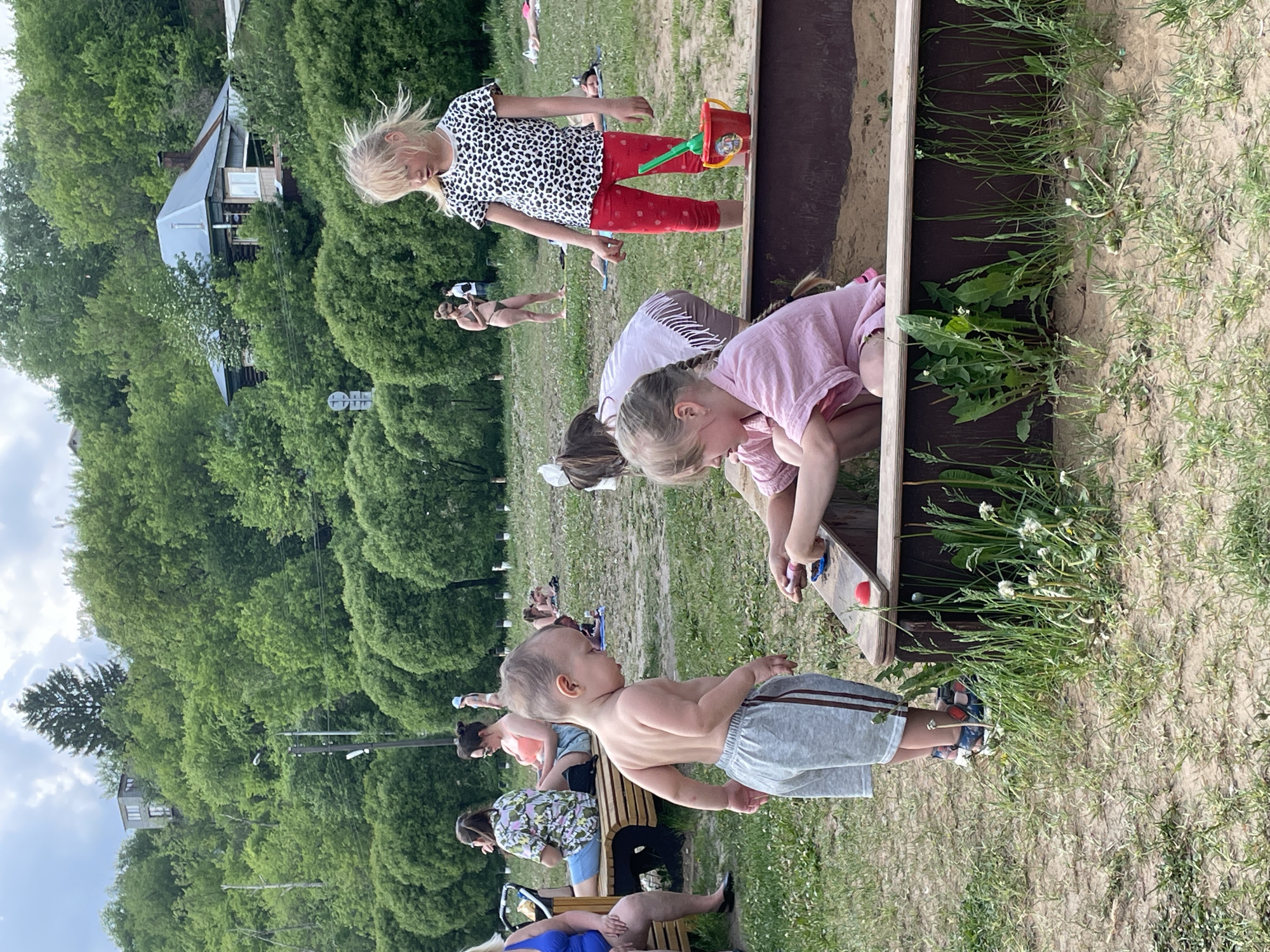 The image shows an outdoor scene, likely a park or recreation area. In the foreground, a toddler stands shirtless in gray shorts and blue sandals near a sandbox. Three girls are clustered around the sandbox, appearing to be playing. One girl wears a spotted top and red leggings and is holding a red bucket with a green shovel. A second girl is in a pink top, leaning over the sandbox. Another child with brown hair is also leaning towards the sandbox. Behind the children, adults are seated on a bench. Further back, people are sunbathing on the grass. The background features green trees and a building. The location is Perm, Russia.