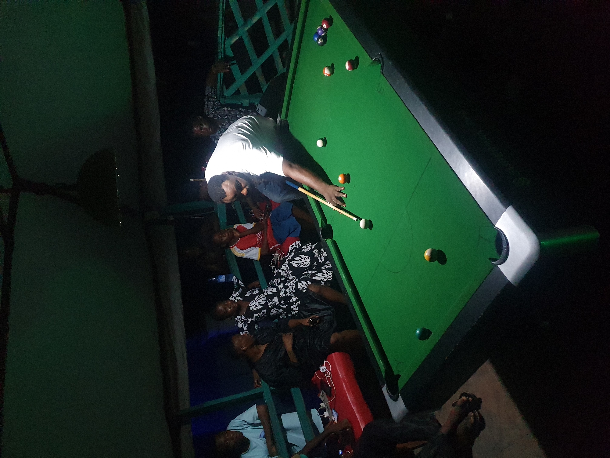 A nighttime scene captures a group of people around a pool table. A man in a white shirt is focused on a shot, cue in hand. Others are gathered nearby, watching or sitting. The green felt of the pool table is visible, with several billiard balls scattered across it. The lighting is dim, but the table is illuminated. The location is Lokoja, Nigeria, indicated by contextual knowledge.
