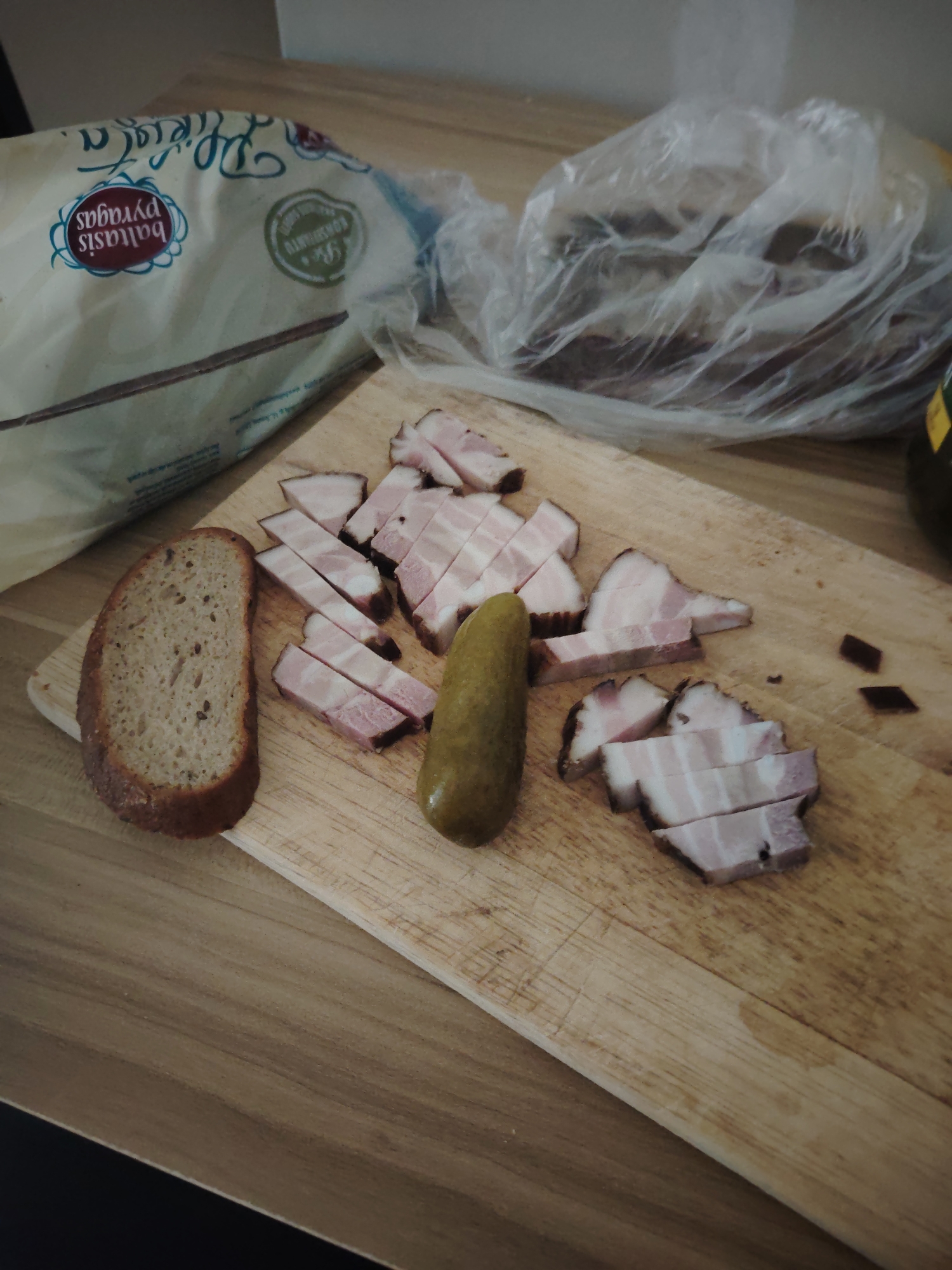 The scene is set in an interior environment in the city of Huntingdonshire, United Kingdom, depicting a selection of food items arranged on a wooden cutting board, which rests on a light wood-grain table surface. Centrally, a rectangular wooden cutting board with visible knife marks holds numerous irregularly sliced pieces of cured or smoked meat, displaying layers of pink flesh and darker, reddish-brown edges. To the left of these meat slices, a single, whole, green pickled gherkin is positioned vertically. Further left, a thick slice of dark, rustic bread with visible grain texture stands upright, partially supported by a smaller wooden piece on the cutting board. In the upper left background, a large, light-colored bag, possibly paper or plastic, partially obscures a loaf of bread and displays text, including "baltasis pyragas" and "pyragas". To the right of the cutting board, a transparent plastic bag contains an obscured dark item, and a small portion of a dark green or brown bottle with a yellow label is visible at the far right edge of the frame. The background consists of a light-colored wall.