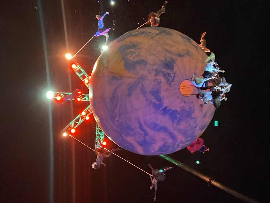 A visually striking scene unfolds in the darkness, featuring a large, globe-like structure painted with Earth's continents and oceans. Acrobats perform on and around the globe, some suspended from ropes extending to a metal structure with lights. A group of people clings to one side of the globe. The overall effect is theatrical, with a focus on aerial performance and a planet Earth theme.