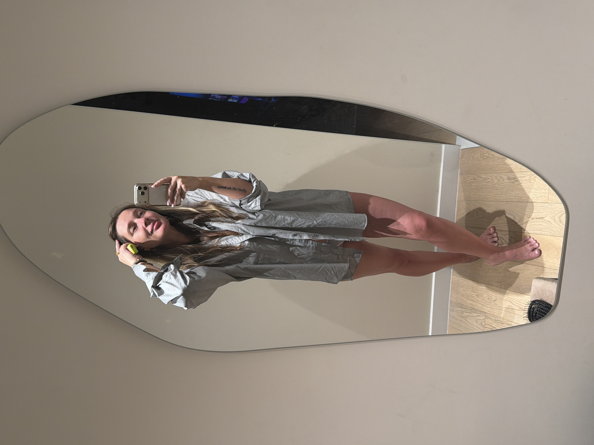 A woman is taking a mirror selfie in what appears to be a dressing room or a similarly well-lit, private space. She is wearing an oversized light blue button-down shirt and is partially visible from the chest down, with her legs and feet in the reflection. She is holding a yellow object in her right hand and has her left hand on her head. The mirror has an irregular, organic shape. The floor is made of light-colored wood planks. There is a black hairbrush with a beige handle visible on a small step in the reflection. The background is a plain, light-colored wall. The image does not provide any specific details that would indicate a location in Moscow, Russia.