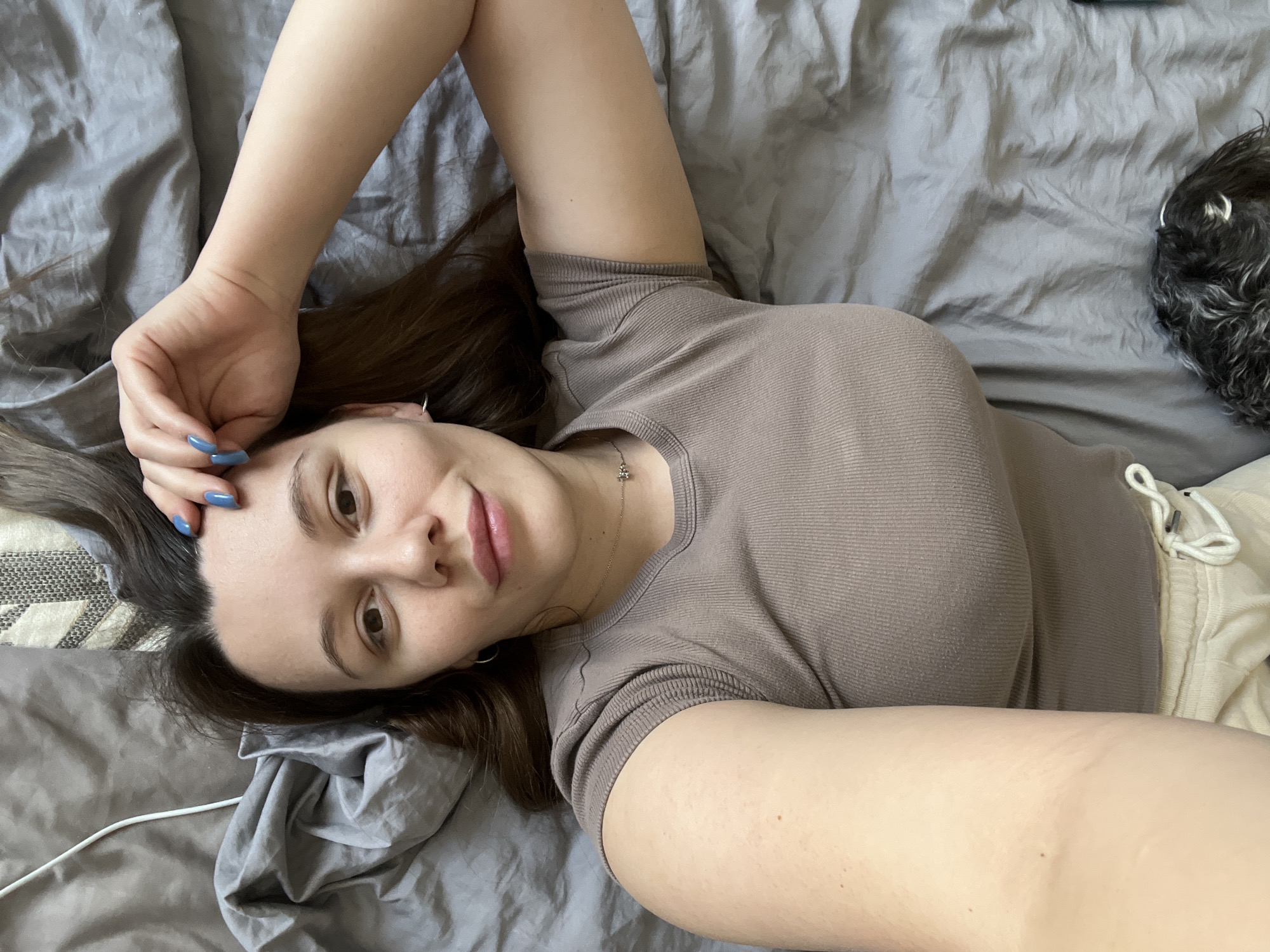 A female subject is depicted lying supine on a bed covered with wrinkled grey sheets. Her head is positioned towards the upper right of the frame, with long, dark brown hair fanned out. She looks directly at the camera with a slight smile. Her left arm is bent, and her hand rests near her forehead, revealing blue fingernail polish. She wears a ribbed, short-sleeved, brownish-grey t-shirt and light-colored (cream) sweatpants or shorts with a visible white drawstring. Accessories include small silver hoop earrings and a thin silver-colored necklace with a small, star-like pendant. A white charging cable is visible on the grey sheets near her head on the left. A small dog, with dark, curly fur mixed with grey, is partially visible at the bottom of the frame, resting on the bed next to her lower body. A portion of a light-colored, patterned headboard or pillow is visible behind her head on the right. The scene is an indoor bedroom setting.