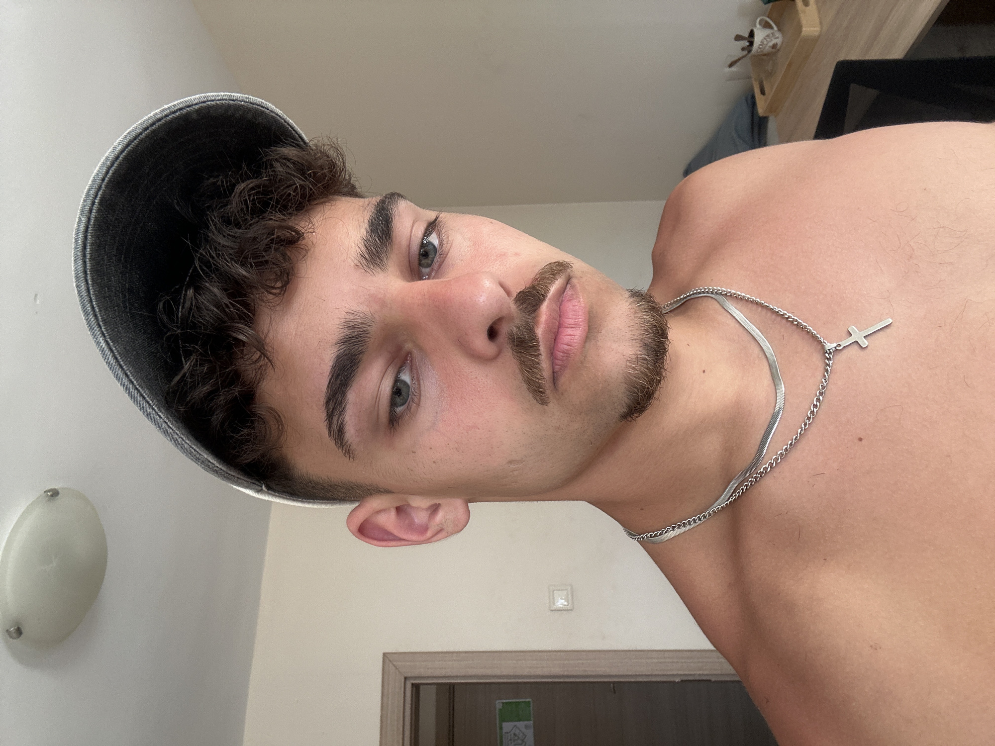 A selfie shot in a room. A young adult male is facing the camera, tilted to the left. He has curly dark hair, gray eyes, and a mustache/beard. He is wearing a denim cap. A silver necklace with a cross pendant is visible around his neck. The background shows a white wall, a light fixture, a door frame, and a partial view of furniture. The location and city is not discernable.