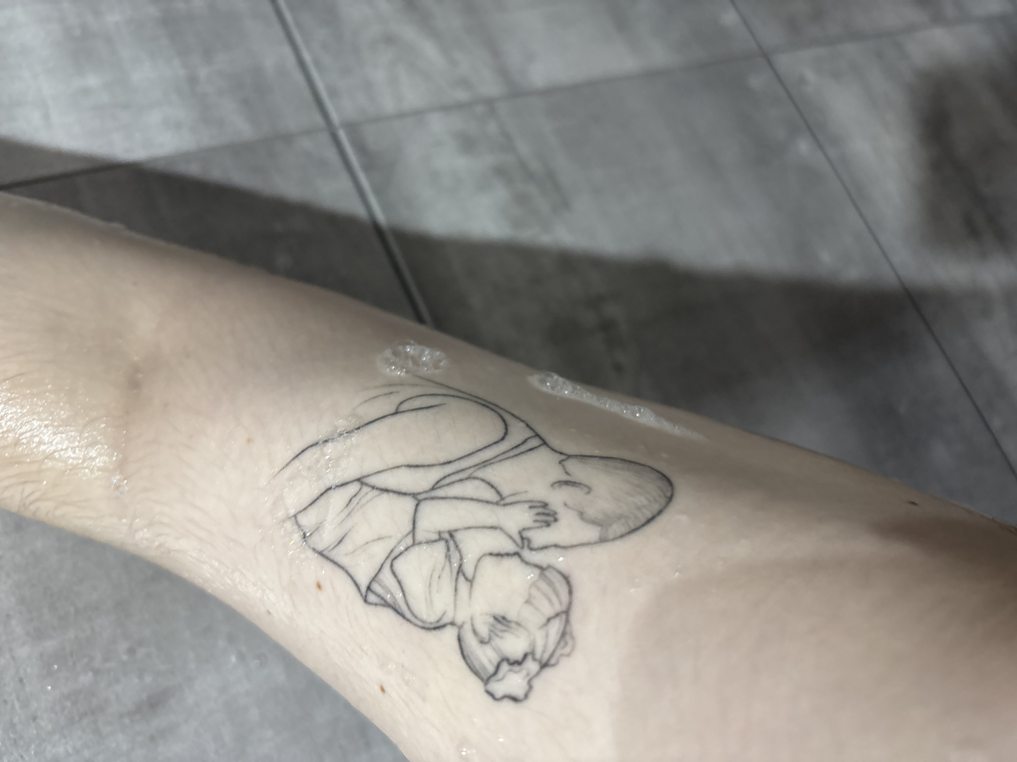 The image displays a human arm, likely a forearm, presented in a vertical orientation. The skin is fair-toned and visibly wet, with numerous small water droplets and bubbles adhering to the surface, indicating recent exposure to water. Fine hair is discernible on the skin. A black outline tattoo is located on the central portion of the arm, depicting two figures in an embrace. One figure, presumably a mother, is shown with an arm wrapped around a smaller figure, presumably a child, whose head rests against the larger figure. The child wears a simple head covering or has styled hair. The background is out of focus, revealing a pattern of grey rectangular tiles with visible grout lines, consistent with an indoor environment such as a bathroom or shower area. The specific location within Perm, Russia, cannot be determined from the visual evidence provided in the image.