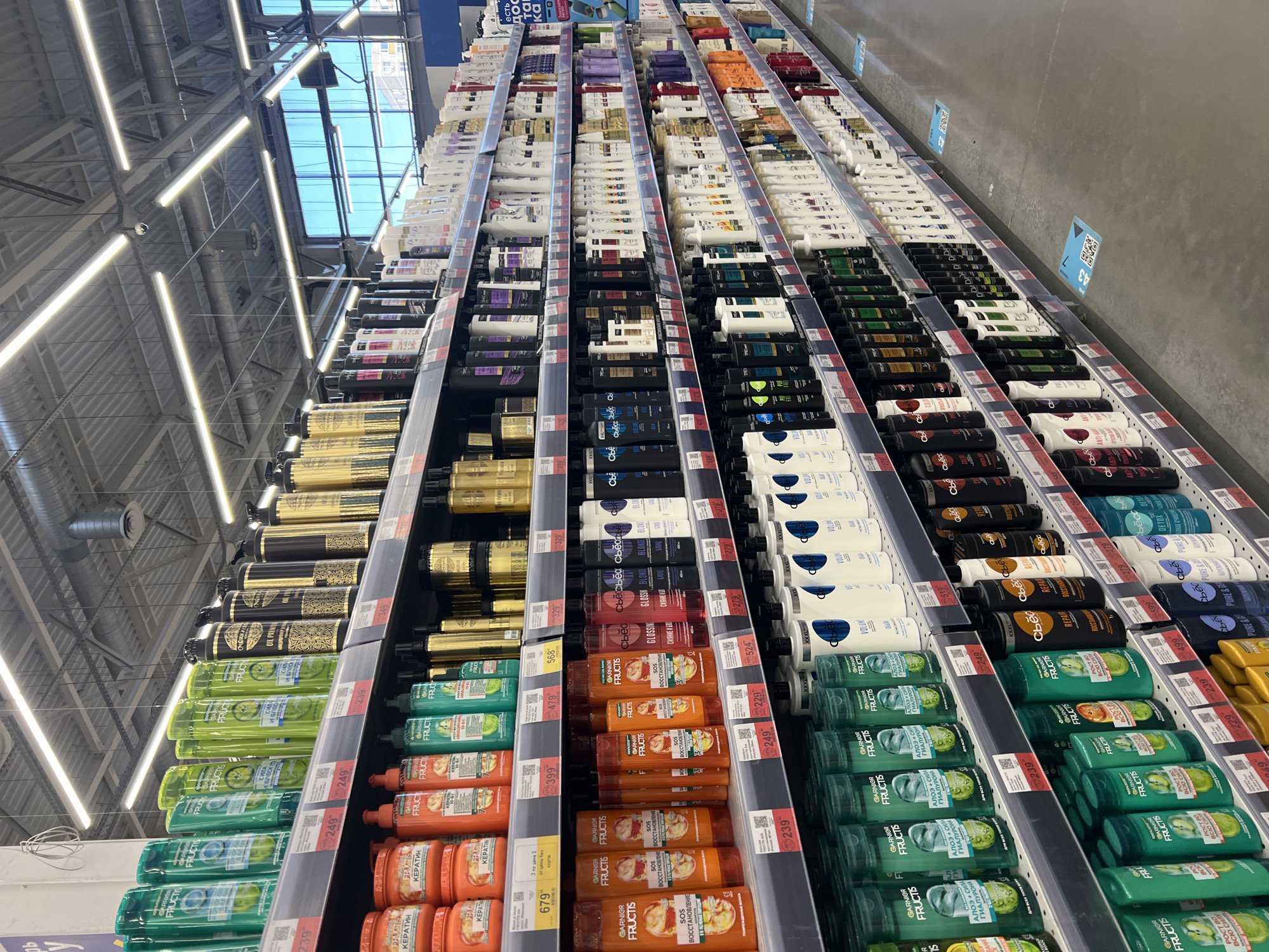 This media file shows a well-lit supermarket aisle stocked with numerous bottles of shampoo and conditioner. The products are arranged on tiered shelving units, with the majority being from the Garnier Fructis line, featuring bright green and orange packaging. Some bottles also display text in Cyrillic, such as "КЕРАТИН" (Keratin), "SOS ВОССТАНОВЛЕНИЕ" (SOS Restoration), and "АЛОЭ + СИЛА" (Aloe + Strength). Price tags are visible on the shelves, with prices like 239, 249, and 679. The lighting suggests an indoor setting, possibly during the daytime due to the bright, even illumination, and the reflection of sky and buildings visible through a skylight in the ceiling. The overall impression is a clean and organized retail environment. There are no people visible in the image. The scene captures a typical retail display of personal care products. The text on the products and price tags provides specific details about the items being sold, and the Cyrillic script indicates a Russian-speaking market, with Krasnodar, Russia specified as the location context.