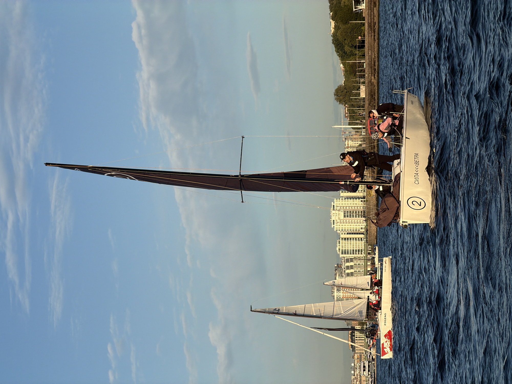 Two sailboats are navigating a body of water with a city skyline in the background. The sailboat in the foreground is white with a dark sail. A person in a dark jacket and cap is standing at the helm, adjusting a rope. Several other individuals are visible on the deck of this sailboat, some wearing life vests. The hull of this boat features the text "СИЛА ««ВЕТРА»" and the number "2" is visible on the stern.
Behind it, another sailboat with white sails is also underway. This sailboat has red and white markings on its hull and displays the word "ZIGZAG". People wearing red life vests are visible on its deck.
In the background, a line of modern buildings with a light-colored facade is situated along the waterfront. Trees and a fence are visible between the water and the buildings. The sky is a clear blue with scattered white clouds. The water appears choppy with moderate waves. The scene suggests a sailing event or recreational activity in Saint Petersburg, Russia, likely on the Neva River or one of its adjacent waterways, given the architecture typical of the city.