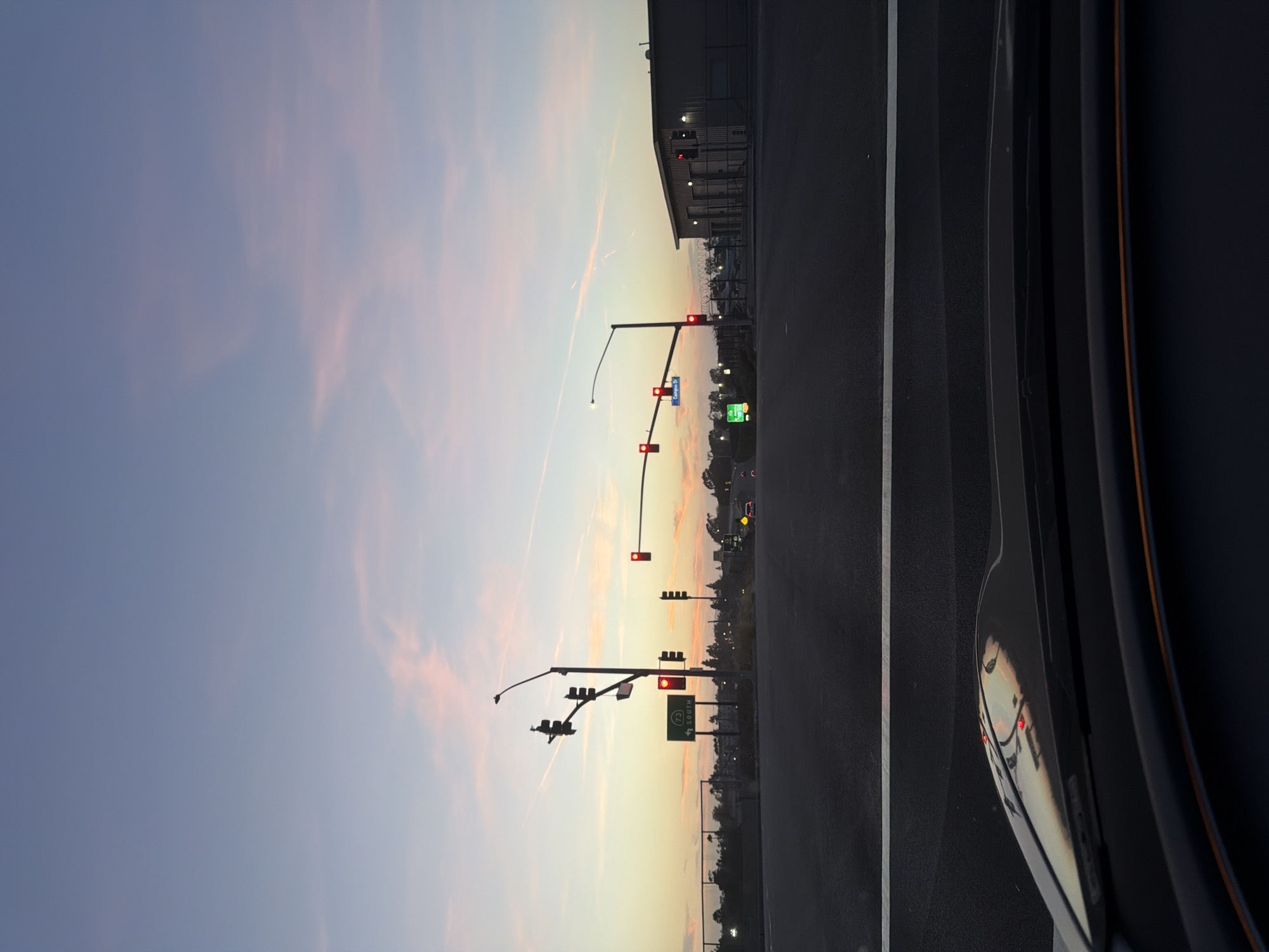The image depicts a sunset scene at a street intersection in Newport Beach, California, United States. The sky is a gradient of pale blue and pink hues, with thin, wispy clouds. In the foreground, the dashboard of a vehicle is visible, reflecting the sky and traffic lights.
The intersection features multiple traffic signals, all illuminated red, indicating that vehicles must stop. A green highway sign with the number "73" and the word "SOUTH" is visible on the left side of the road. A blue street sign reading "Campus Dr" is also present. In the background, buildings and streetlights are silhouetted against the twilight sky. A chain-link fence runs along the side of the road. The road surface is asphalt, with a white lane marker clearly visible. No people are visible in the frame.