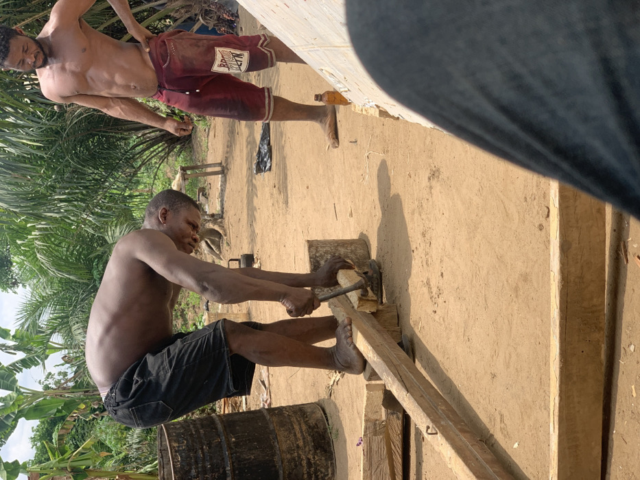 The image captures two men in an outdoor setting. One man, shirtless, is wearing red shorts with the inscription "Brown Bell" visible. He stands to the left of the frame and looks towards the center. The other man, also shirtless, is wearing black shorts. He is positioned in the center of the frame, crouching down and using a metal tool to work on a wooden plank. The plank is resting on two other pieces of wood, and there is a piece of wood beneath it. The ground appears to be loose dirt, and there are various pieces of wood scattered around. Behind the men, there are lush green plants growing in the background. The location is Egbeda, Nigeria, based on the provided coordinates (latitude: 7.12, longitude: 5.362).