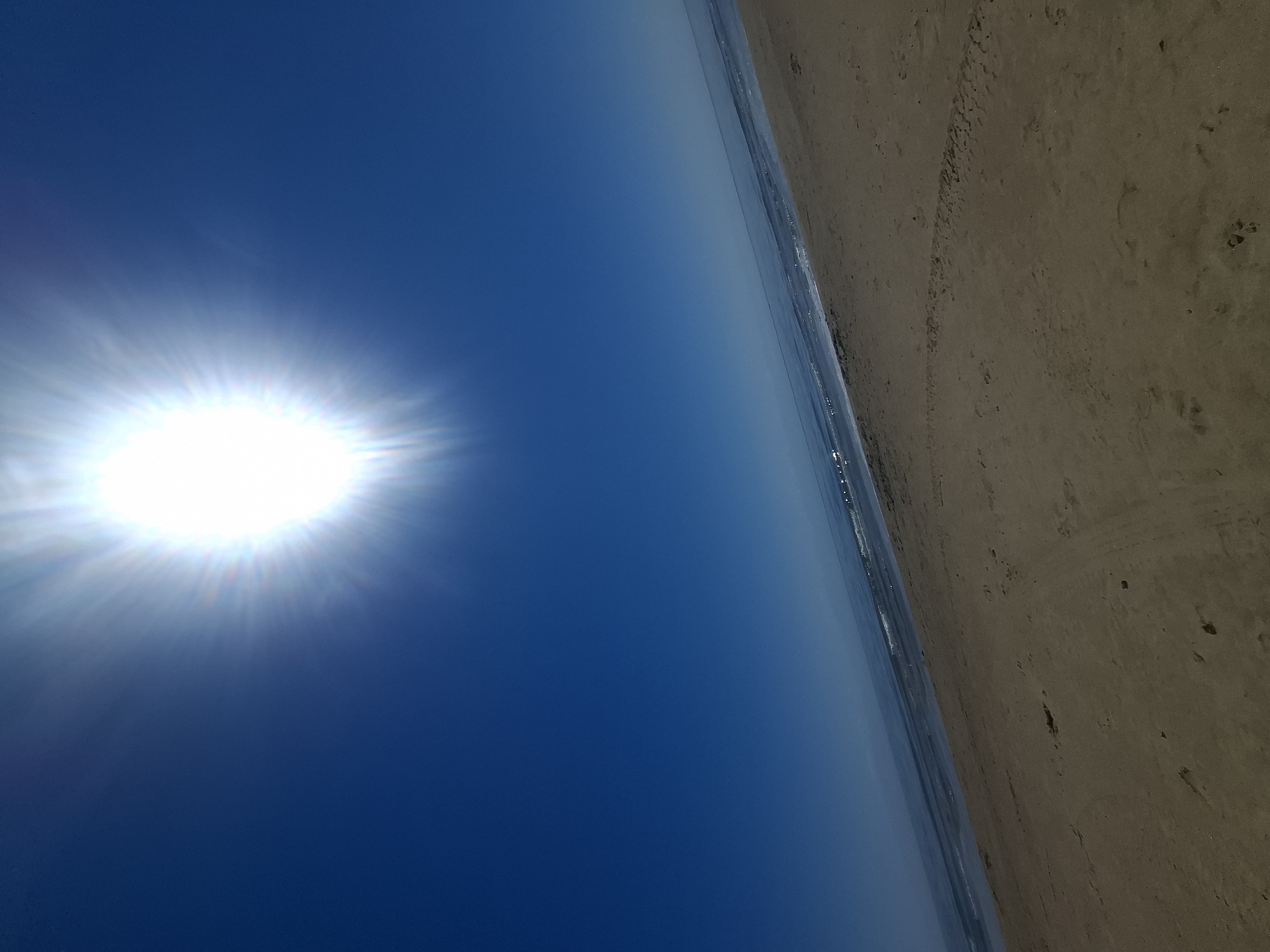 The image shows a wide shot of a sandy beach with the sun shining brightly in a clear blue sky. The sun is positioned in the upper center of the frame, casting rays of light. The ocean is visible in the background, with gentle waves lapping at the shore. The sand is a light brown color, and there are some footprints visible on the beach. The scene appears to be a sunny day at the beach. The location is specified as the city of Qonaev, Kazakhstan.