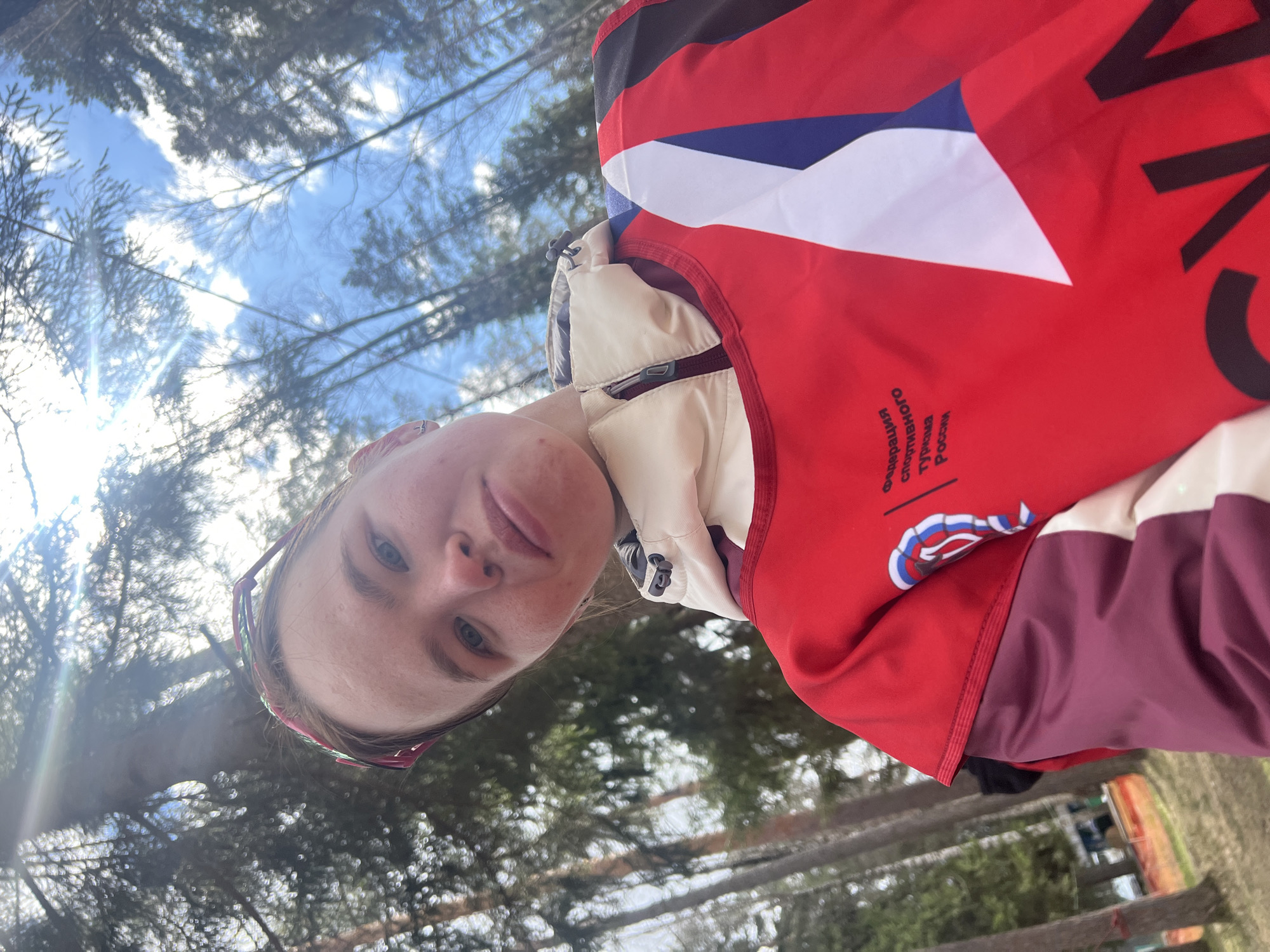 A young woman is holding a camera and taking a selfie. She is outside in a wooded area on a sunny day. The woman is wearing a white jacket and a red sports vest that says "Федерация спортивного туризма России," indicating she may be involved in sports tourism in Russia. Based on the vest, the location might be Lesnoye, Russia. The background features trees and a blue sky with some clouds.