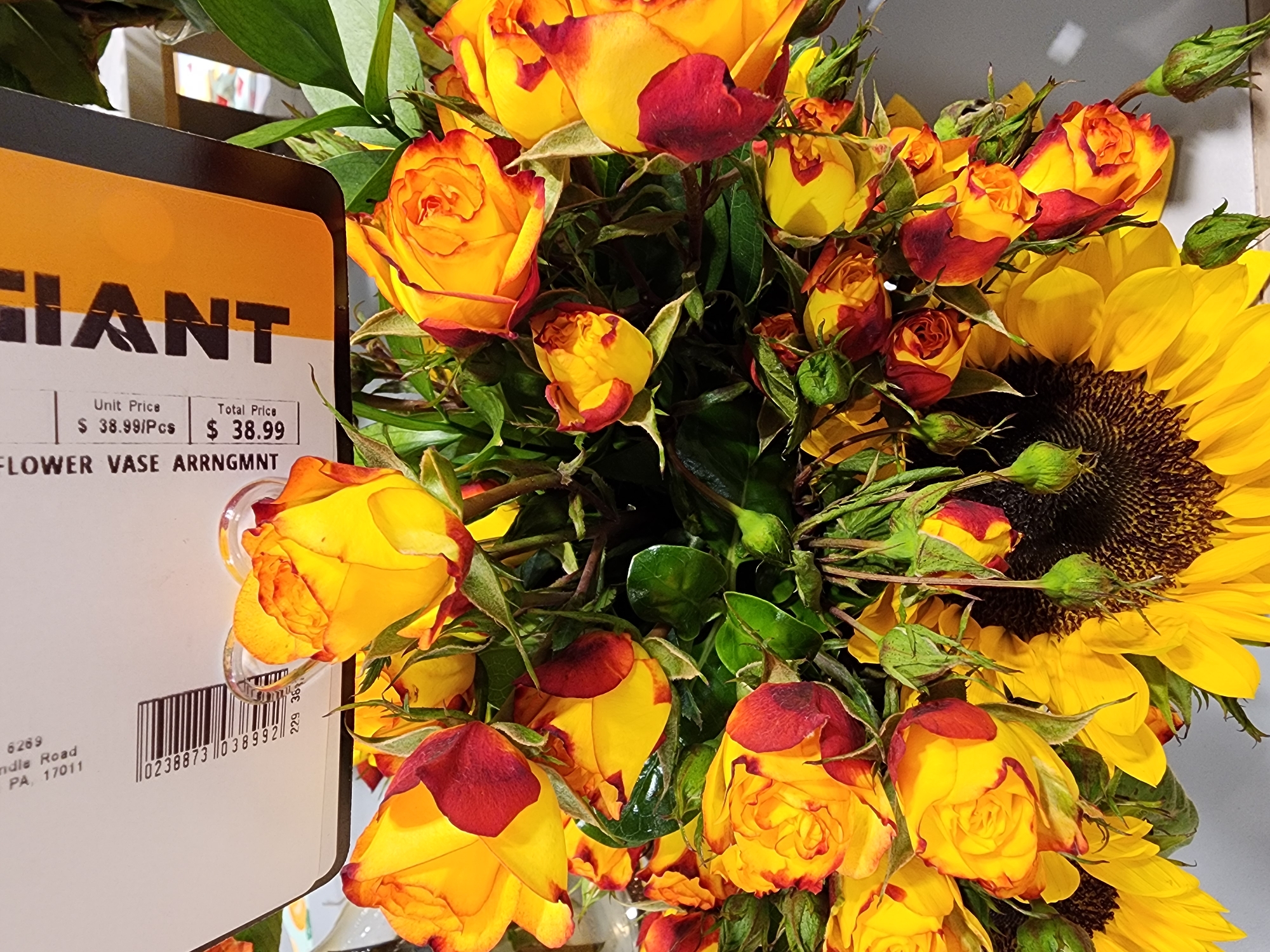 The image shows a bouquet of flowers, predominantly yellow roses with red-tipped petals and sunflowers, arranged in a clear vase. The bouquet is displayed against a plain background, with a price tag attached to the front of the vase. The price tag includes information such as "FLOWER VASE ARRNGMNT," "Unit Price $38.99/Pcs," and "Total Price $38.99." There is also a barcode and an address listed on the tag: "6269 Candle Road, Camp Hill, PA, 17011." The lighting suggests an indoor setting, likely a retail environment.