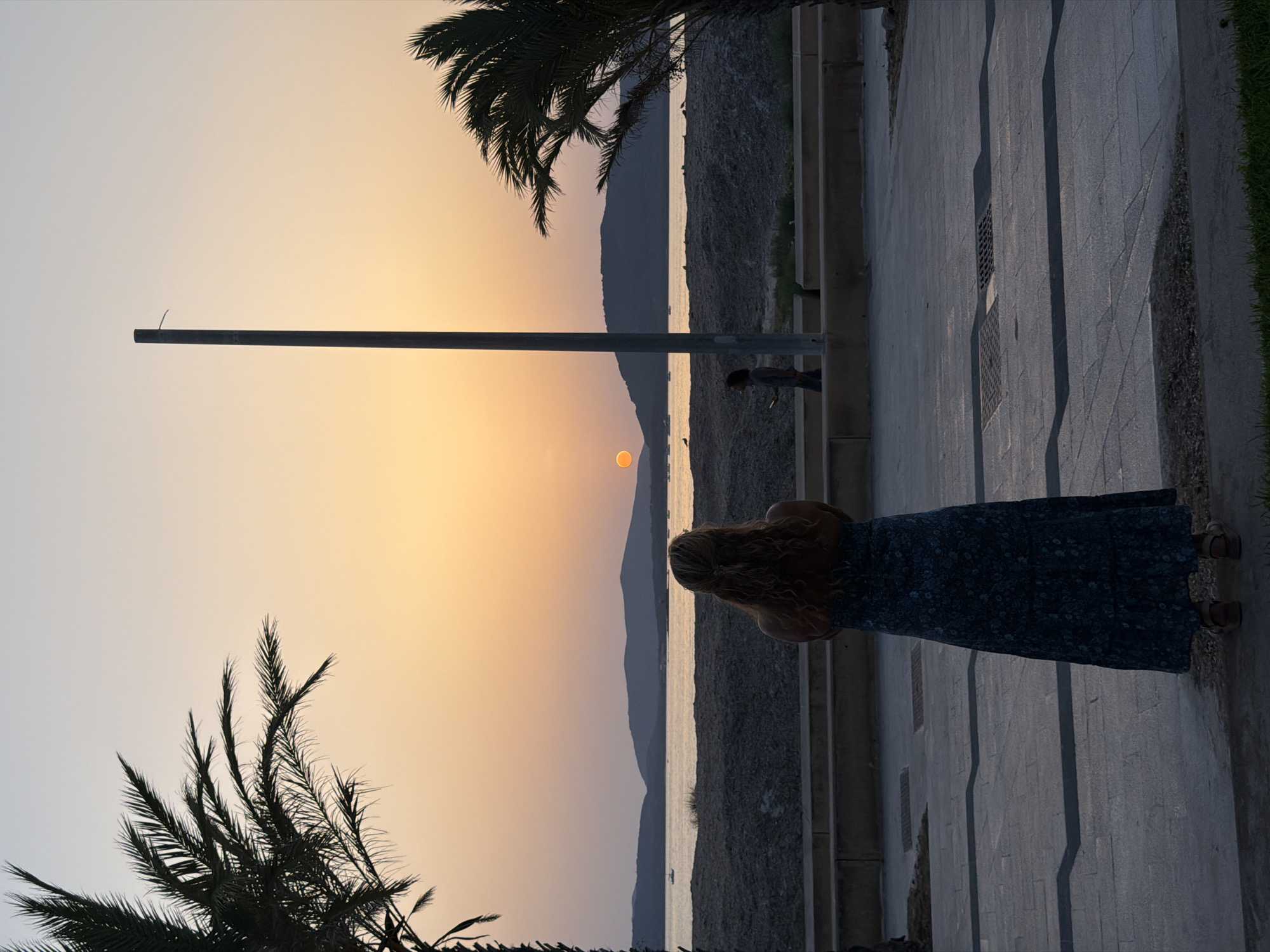 The image depicts a sunset scene by the sea. In the foreground, a woman with long, wavy blonde hair is seen from behind, wearing a dark, floral-patterned maxi dress and sandals. She is standing on a paved area with geometric patterns. To her left, a palm tree's silhouette is visible. In the midground, a beach with dark sand and rocks separates the paved area from the sea. A low concrete barrier runs along the edge of the paved area. Further back, a body of water is visible with several small boats. In the distance, dark, silhouetted hills form the horizon. The sun, a bright orange orb, is setting over the sea, casting a warm glow across the sky. A tall, thin pole stands vertically in the midground. Another person, silhouetted, is walking in the background. The overall setting suggests a coastal promenade or walkway. The image captures a serene moment during a sunset. This scene is evocative of the waterfront areas of Alghero, Italy, known for its picturesque sunsets over the Mediterranean Sea.