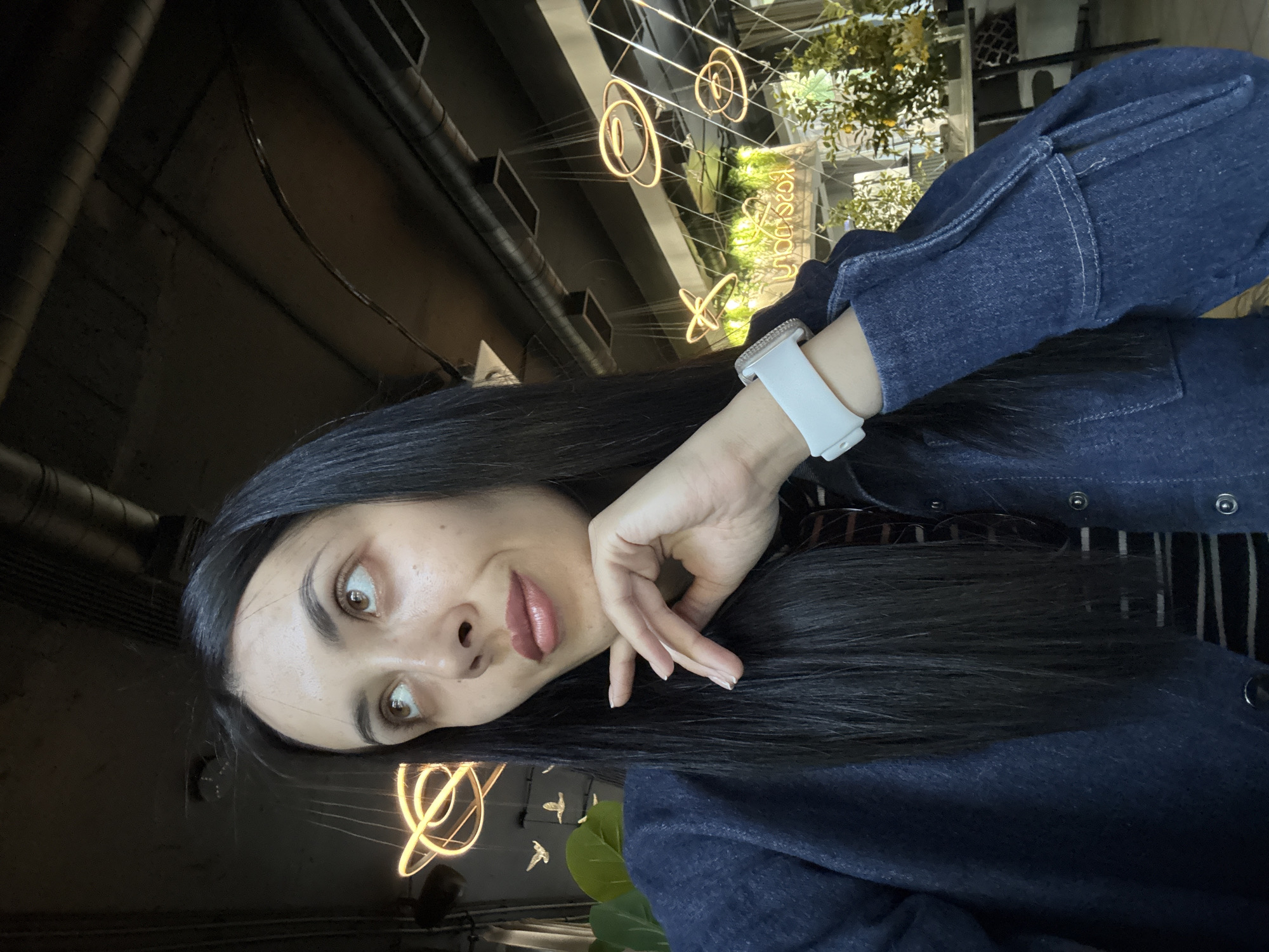 The image shows a woman with long, dark hair wearing a dark blue denim jacket and a white smartwatch. She is gazing at the camera with a slightly inquisitive expression, her hand near her face. The background is a dimly lit interior with exposed ceiling pipes and modern light fixtures shaped like rings and butterflies. Through a window, there are glimpses of greenery and what appears to be a sign in Russian. The location cannot be definitively identified as Votkinsk, Russia, as the available visual details are not sufficient for specific geographic determination.
