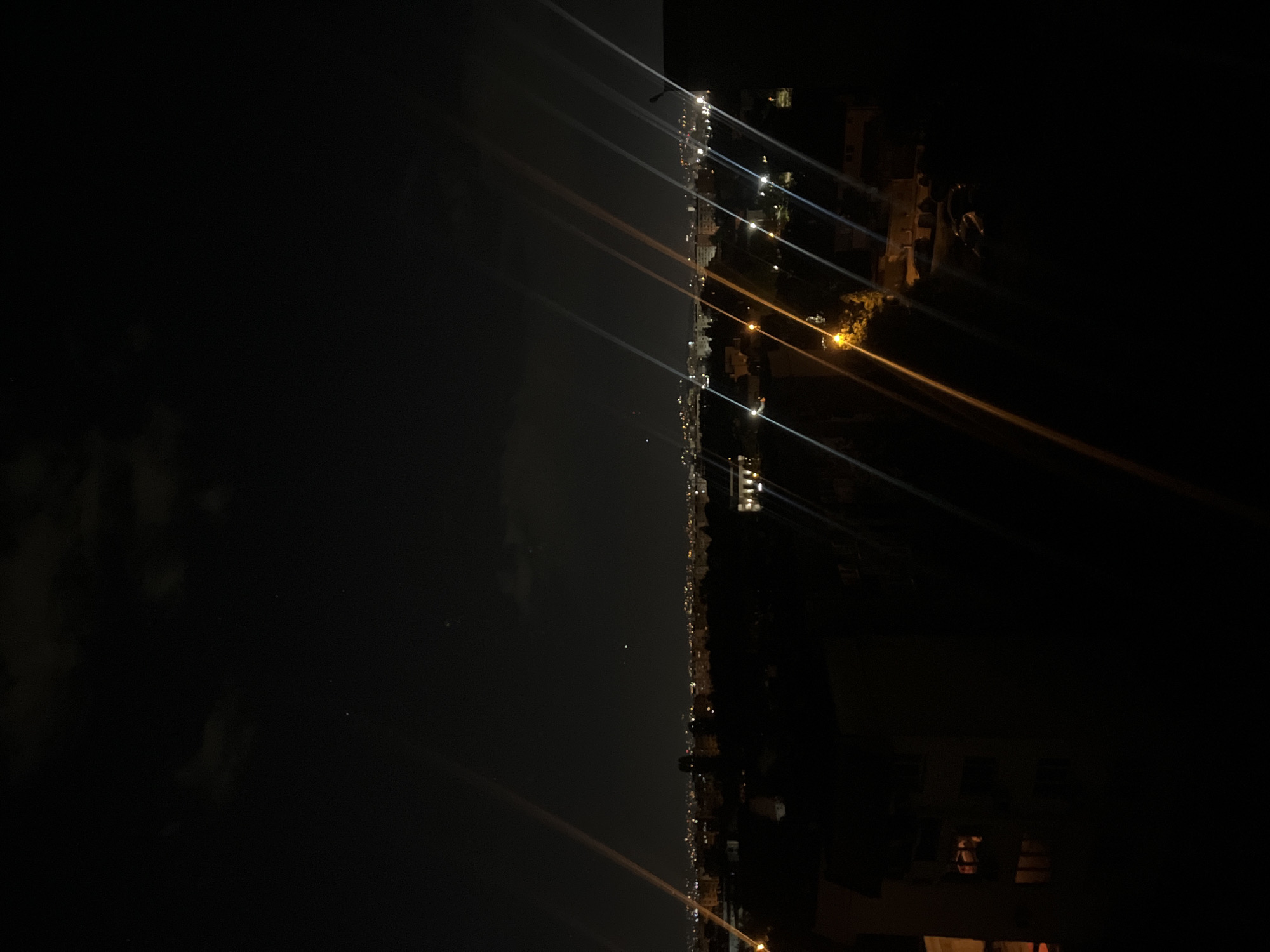 A nighttime scene shows the city of Sofia, Bulgaria, as viewed from a high vantage point. Illuminated buildings and streetlights trace the cityscape, forming a line between the dark sky on the left and the darker urban environment on the right. Light beams radiate upwards from the streetlights, creating a series of streaks against the night sky. Individual buildings and vegetation are discernible despite the darkness. The image is oriented sideways.