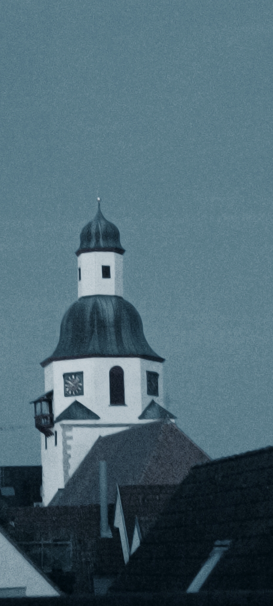 The image is a slightly grainy, vertically oriented photograph of the tower of the Evangelische Stadtkirche (Evangelical City Church) in Winnenden, Germany. The church tower is predominantly white with a dark, domed roof topped with a small spire. On the tower, there is a clock face with Roman numerals and hands indicating approximately 10:08. There is also a balcony-like structure protruding from the tower. Several dark-colored rooftops of other buildings are visible in the foreground, obscuring the base of the church tower. The sky is a uniform, overcast blue-gray. There are no people or animals visible in the image. The overall lighting suggests it is daytime, but the sky is heavily clouded.