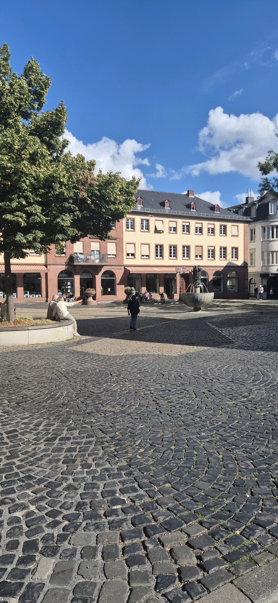 This is a vertical, eye-level shot of a cobblestone plaza in Mainz, Germany, under a bright blue sky dotted with fluffy white clouds. The plaza is lined with traditional European buildings featuring cream-colored facades, dark tiled roofs, and red-trimmed dormer windows. In the foreground, the cobblestone pavement is laid in concentric circles, with a large tree on the left casting a shadow.
To the left, a person in a light-colored shirt and pants sits on a low stone wall, holding a phone and appearing to take a selfie. A person in a dark jacket and jeans walks away from the camera towards the center of the plaza. In the mid-ground, a large, ornate fountain with multiple tiers is active, with water flowing from the top. Behind the fountain, the buildings have arched ground-floor entrances leading to shops. Several people are visible in the distance, seated at tables outside a cafe or walking past the shops. The overall atmosphere is bright and sunny, suggesting a pleasant day in a European city square.