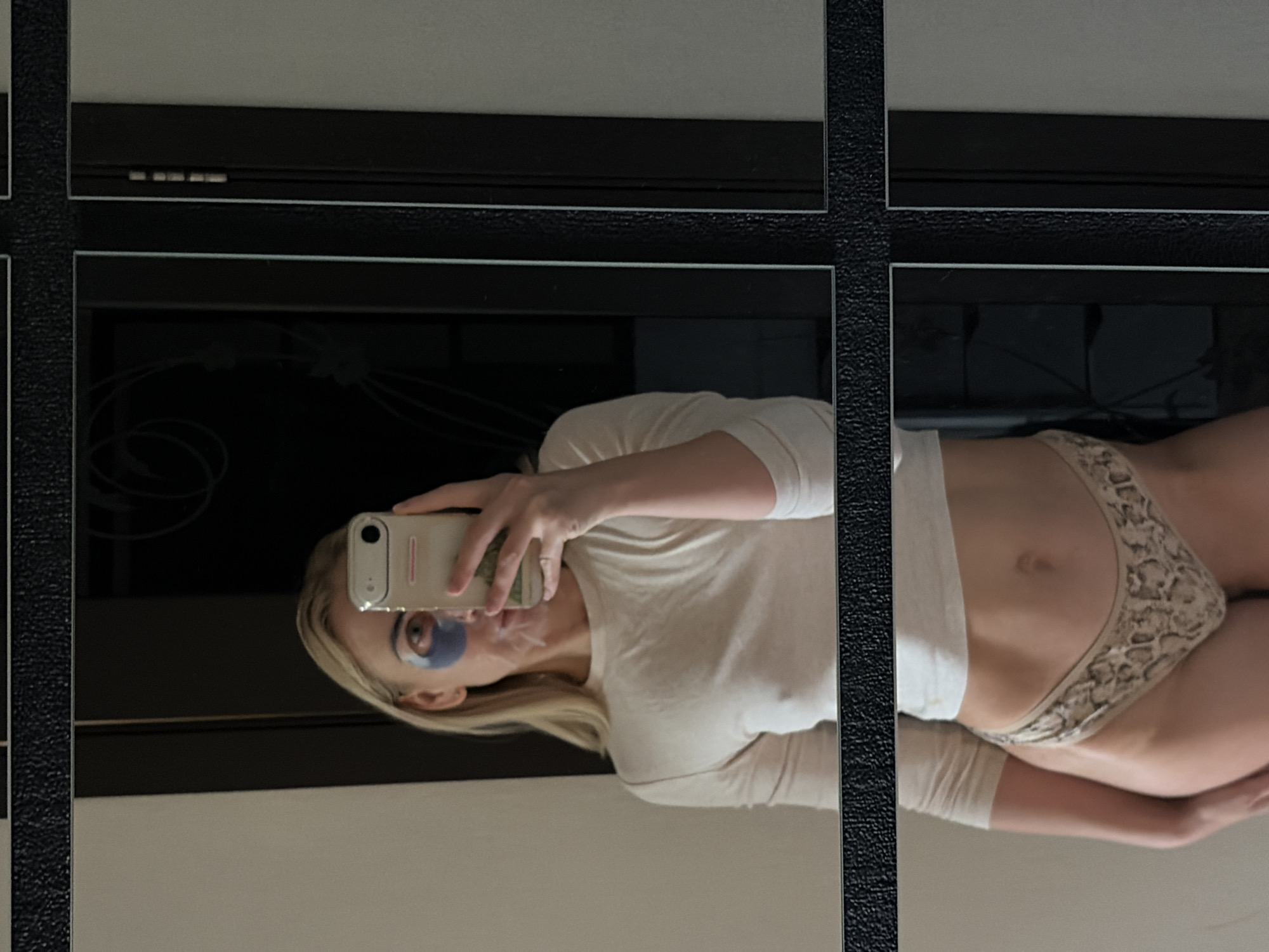 A blonde woman with blue under-eye patches is taking a mirror selfie. She is wearing a light-colored long-sleeved shirt and patterned underwear. The reflection shows her face and upper body in the top half of the image, and her lower body in the bottom half. The background appears to be a room with mirrors and dark doorways. The image quality is moderate, with some graininess. There is no information in the image to suggest the location is Chaikovsky, Russia.