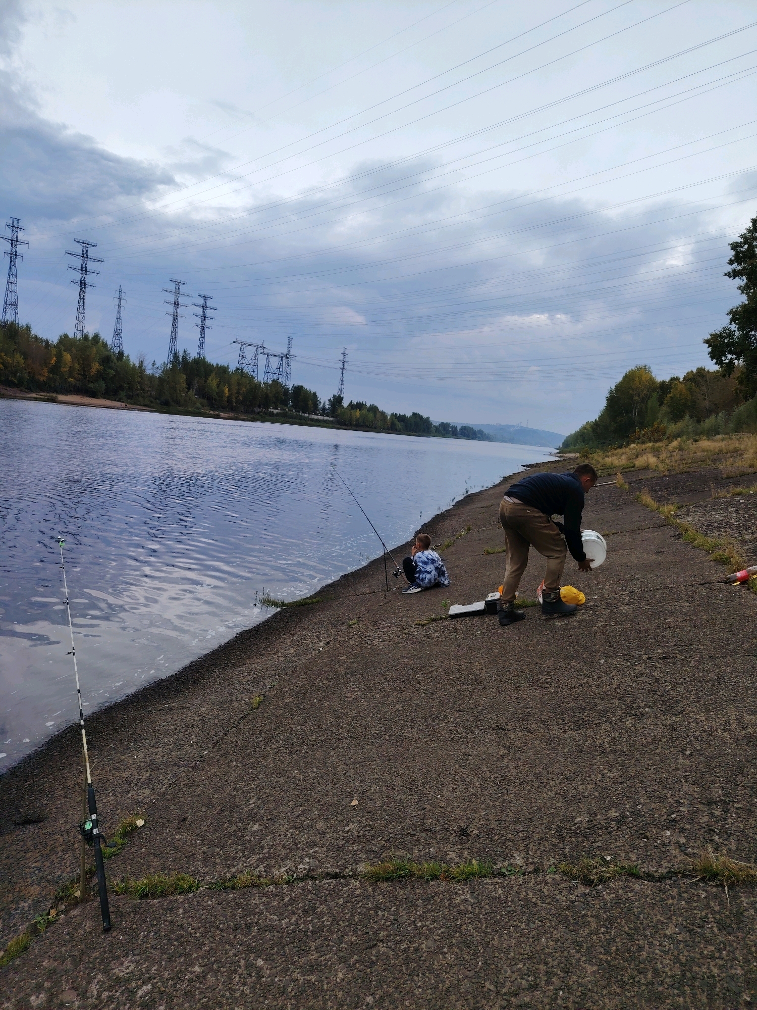 The image depicts a view of the Kama River embankment in Chaikovsky, Russia, on a cloudy day. In the foreground, a concrete embankment runs along the river. Two fishing rods are visible, one in the lower left of the frame partially submerged in the water, and another standing upright further down the embankment on the right.
On the embankment, a man wearing a dark long-sleeved shirt and brown pants is bent over, holding a white bucket in his right hand and looking down. Near him, a yellow object, possibly a bucket lid or another container, lies on the ground. A young child, wearing a blue and white tie-dye shirt, is sitting on the embankment facing the river, a short distance from the man.
In the background, the Kama River is calm, with ripples on its surface. The opposite bank of the river is lined with trees exhibiting autumn colors. In the far distance, power transmission towers and lines are visible against the overcast sky. The overall scene suggests a recreational fishing activity.