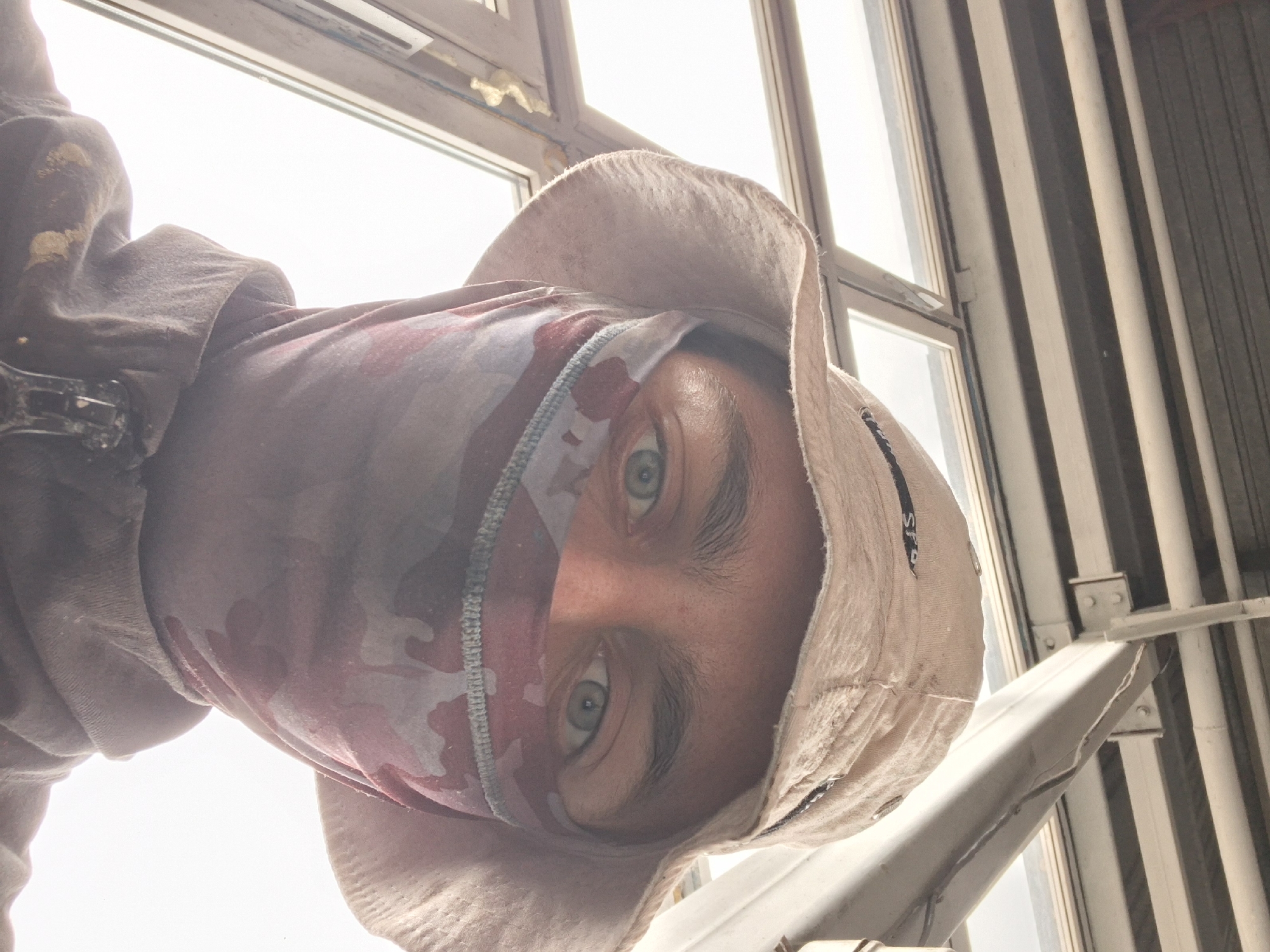 A person is wearing a hat and face covering. The individual has light skin, visible eyes, and eyebrows. The hat is light brown, while the face covering is red and white camouflage. The person is positioned indoors, near a window with a white frame. Vertical pipes and metal supports are visible behind them. The location and city are unknown.