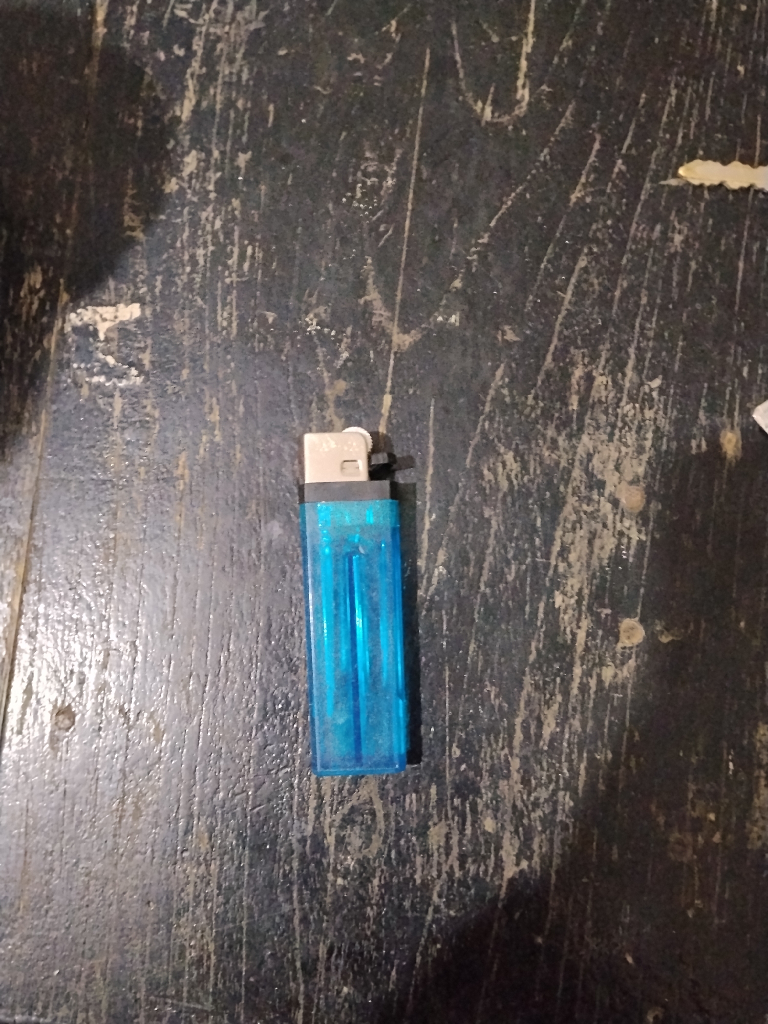 The image shows a blue disposable lighter lying on a dark, textured surface that appears to be wood, possibly a table or floor. The surface has visible scratches and imperfections, suggesting age or heavy use. The lighter is oriented vertically and is the central subject of the image. There are no people or other objects clearly visible in the frame. The lighting is dim, casting subtle shadows. The scene is devoid of any specific context that would suggest a location in Bilis Bilis, Indonesia, or any other specific geographical area.