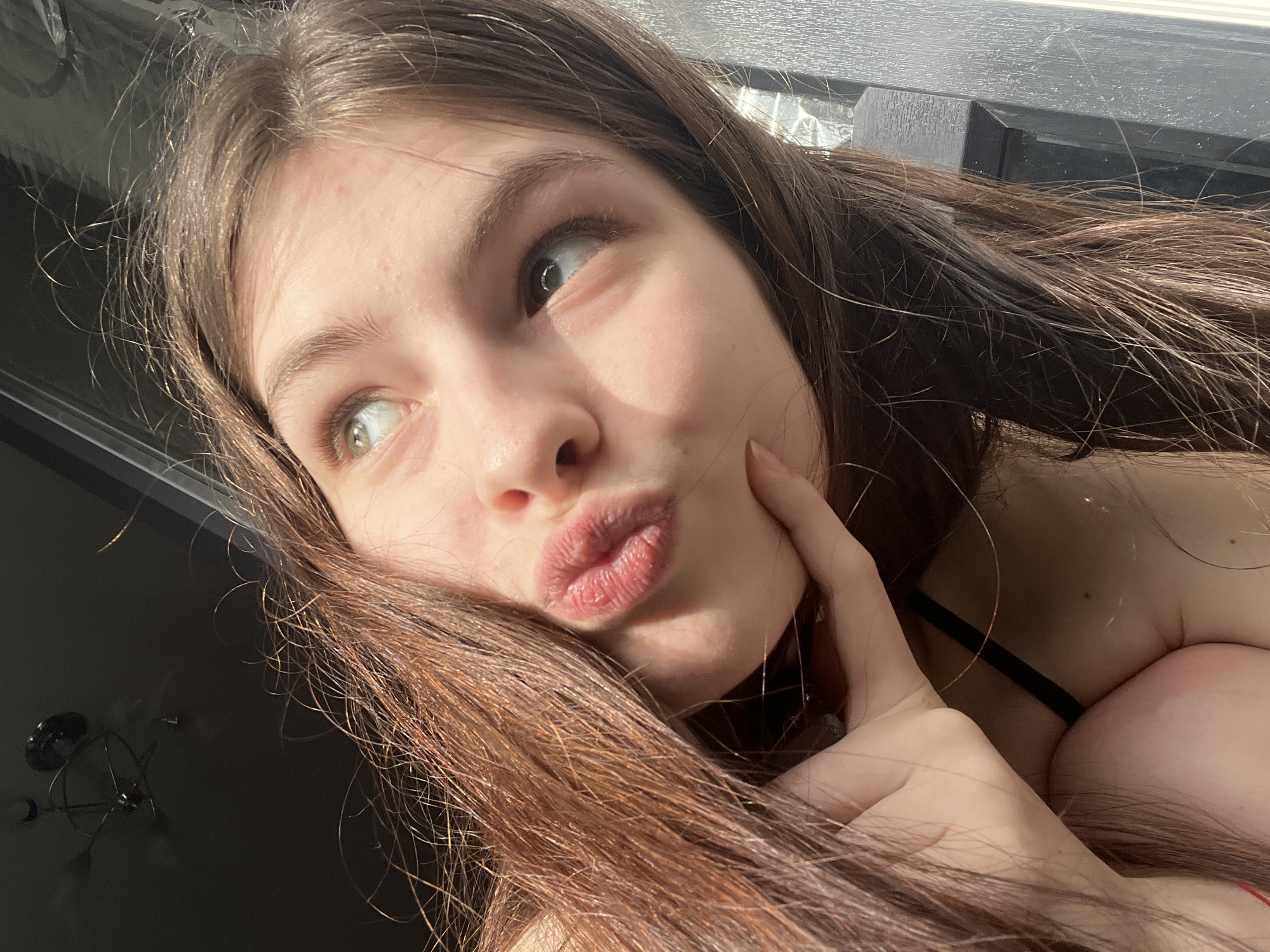 A young woman with long brown hair is posing for a selfie, puckering her lips. She has fair skin and her hand is touching her cheek. A black bra strap is visible. The background includes a window frame. The location is unknown and there is no indication that it is in Saint Petersburg, Russia.