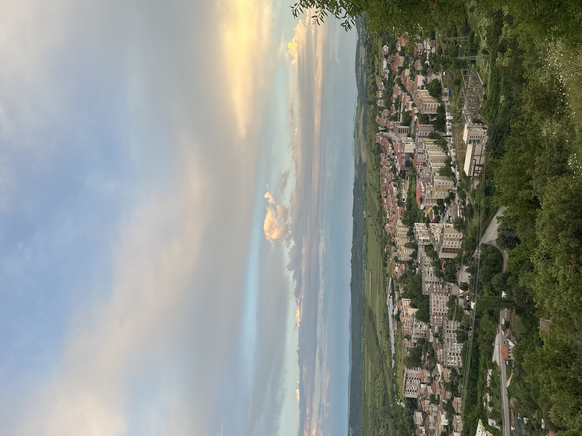 The image shows an aerial view of Ivaylovgrad, Bulgaria. The right side depicts residential buildings and greenery, blending into a forested area. Electrical power lines are visible. The left side features a sky filled with clouds, tinged with shades of yellow and blue.