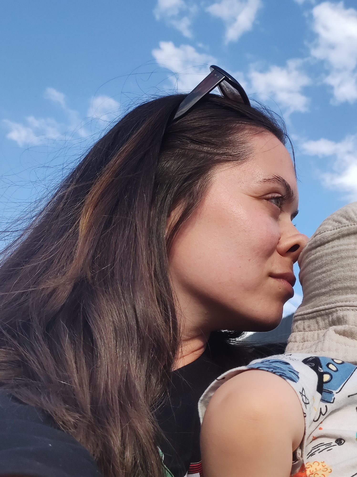 A medium shot presents a woman with long, dark hair and sunglasses positioned atop her head, captured in profile against a backdrop of a blue sky dotted with scattered clouds. Her gaze is directed towards the right of the frame, where a child's hooded head and clothed shoulder are visible. The woman's skin is fair, and her facial features are clearly defined in the outdoor lighting. The child's clothing features a cartoon-like design. The scene appears to be taking place outdoors, possibly in Chaikovsky, Russia, but there are no specific landmarks visible in the shot to definitively confirm the location.