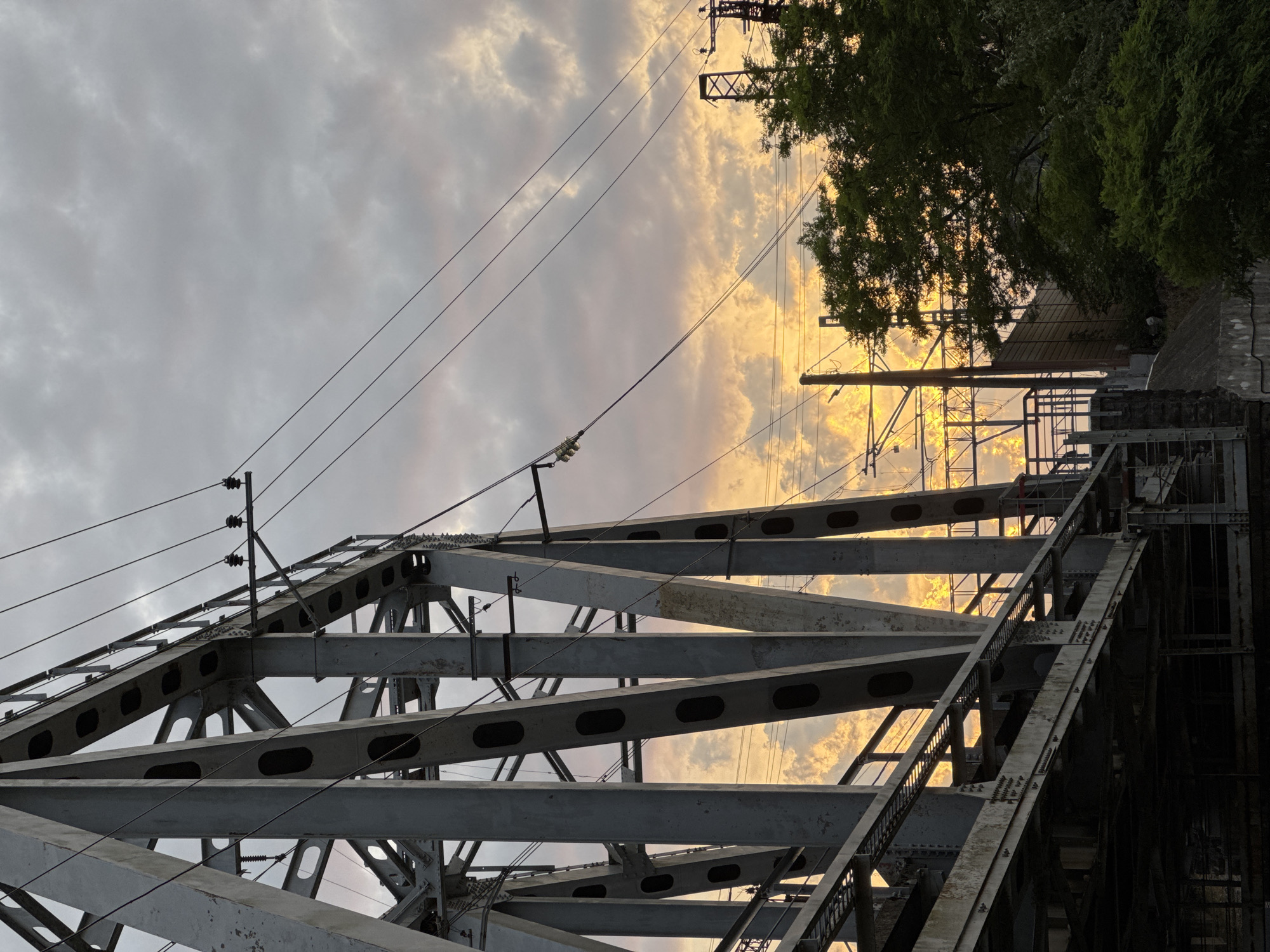 The image shows a view of a railway bridge at dusk. The bridge is made of steel girders and has overhead power lines. The sky is cloudy with dramatic orange and yellow hues from the setting sun. To the right of the bridge, there are trees and some industrial structures. The scene is captured from a low angle, looking up at the bridge and sky. The location is identified as Uch-Dere, Russia.