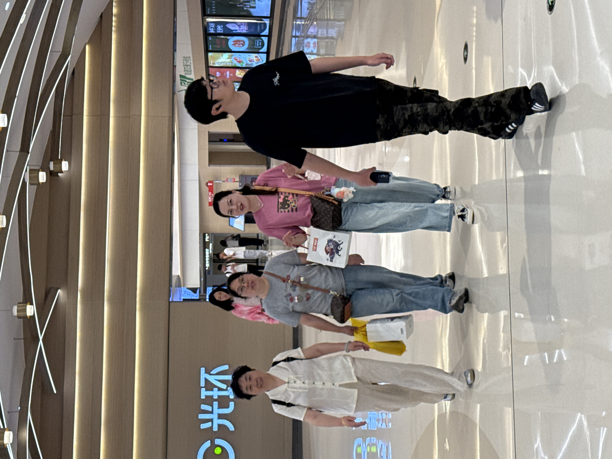The image shows a group of people walking in an indoor setting, likely a shopping mall or commercial space, located in Liangjiang, China. The individuals appear to be shopping or passing through the area.
The people, from top to bottom of the image, consist of: a young man in a black shirt and camouflage pants; a young woman in a pink shirt and jeans, carrying a brown purse; an older woman in a gray shirt and jeans, carrying shopping bags; and an older woman wearing a white blouse and pants.
The environment is modern, with glossy flooring and walls decorated with vertical illuminated panels. Chinese characters are visible on a wall, possibly signage for a shopping center. The general atmosphere suggests a contemporary urban setting.
