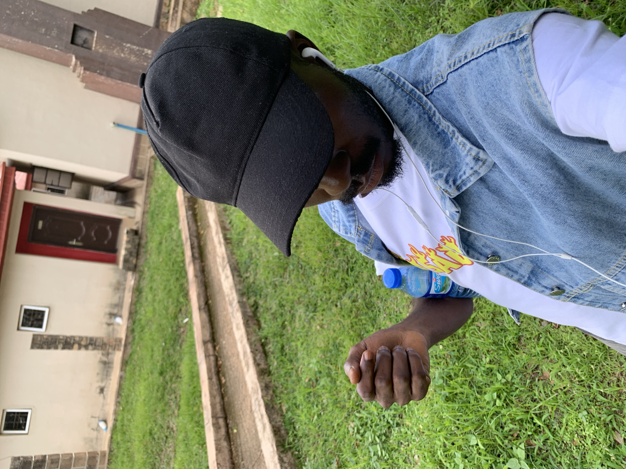 A person wearing a black baseball cap, a denim vest over a white long-sleeved shirt, and white earphones is shown in a slightly tilted, close-up shot. The person has dark skin and a beard. A water bottle with a blue cap is visible. The background is out of focus, showing green grass and a building with a brown door and two small windows. The building appears to be light-colored with some brown architectural elements. The scene suggests an outdoor setting, likely in a residential or institutional area, given the presence of the building and manicured lawn. The lighting indicates it is daytime. The overall mood seems relaxed, possibly indicating a casual outing or break. No specific activity is clearly depicted, but the person appears to be holding the water bottle. The location context is Jos, Nigeria, which is consistent with the outdoor environment and building style.