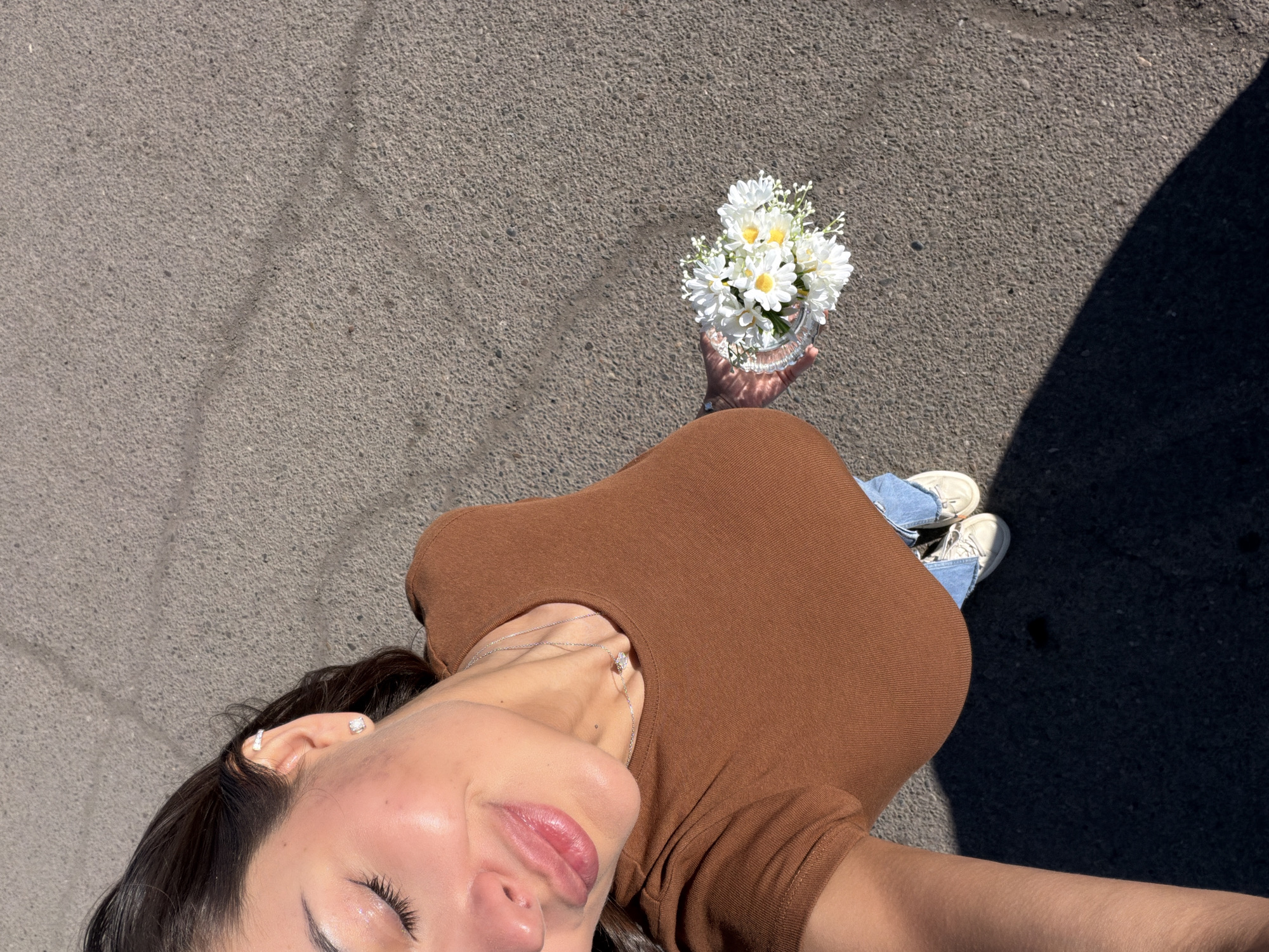 An overhead shot captures a young woman lying or reclining on grey asphalt pavement in Krasnoyarsk, Russia, under bright sunlight. She is seen from the chest up, looking towards the camera, with her eyes closed and a soft smile on her lips, suggesting a moment of peace or enjoyment. Her dark hair frames her face. She wears a fitted, short-sleeved brown top, blue jeans, and white sneakers, partially visible towards the bottom right.
Her right arm is extended upwards, holding a small, clear glass container filled with a bouquet of white daisy-like flowers. The flowers have yellow centers and small green accents. Strong, dark shadows are cast across the asphalt, indicating a sunny day, likely midday or early afternoon. The ground is a uniform texture of coarse asphalt with a few faint lines or cracks. The woman wears small silver stud earrings and a delicate silver necklace with a tiny pendant. No visible text is present in the image.