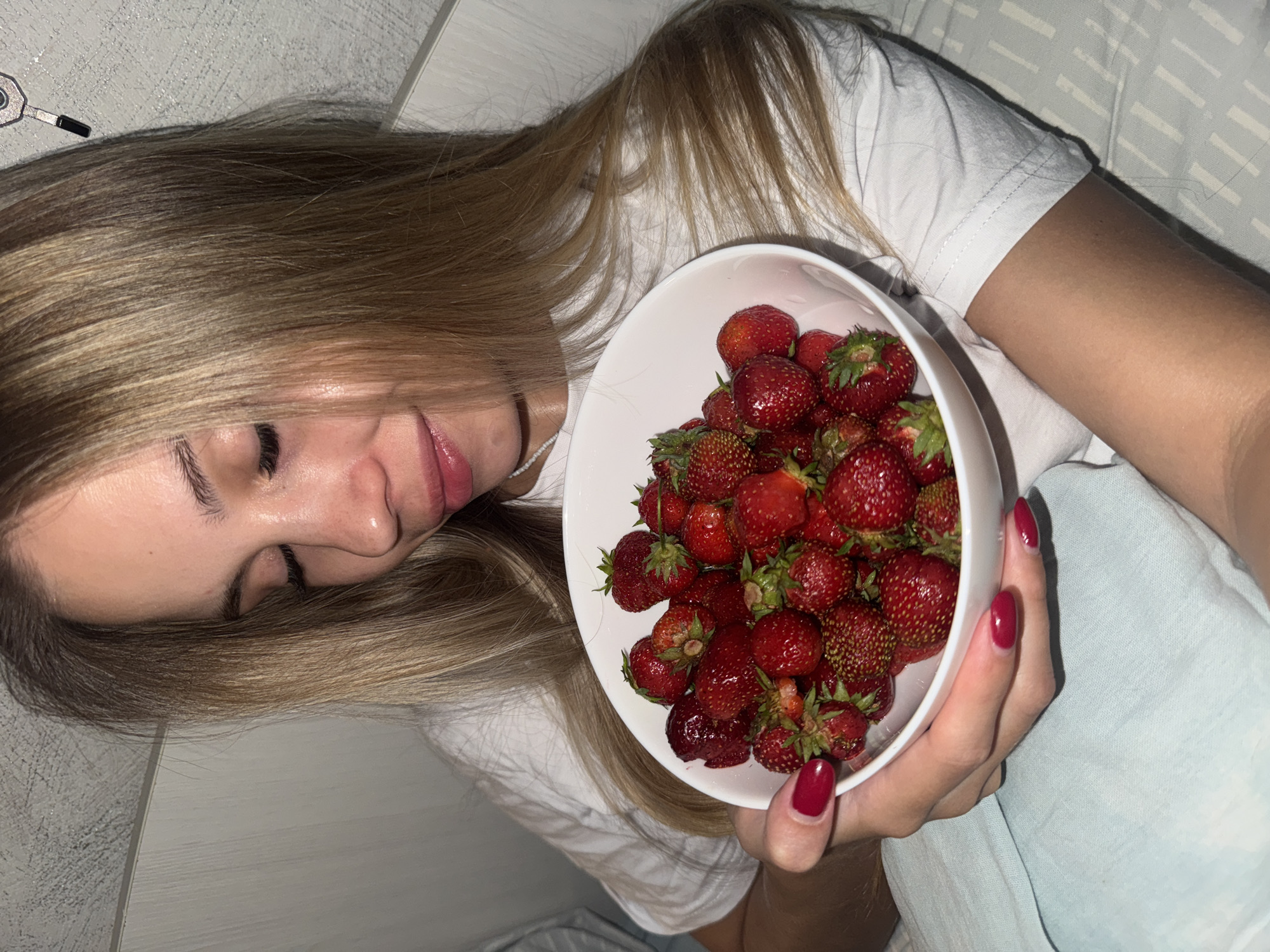 A young woman with blonde hair and red nail polish is lying down, holding a white bowl filled with fresh strawberries. Her eyes are closed, and she has a slight smile on her face, suggesting a relaxed or contented mood. The background appears to be a bed with light-colored bedding, possibly indicating a bedroom setting. The lighting suggests it might be nighttime or an indoor setting with artificial light, as there are no strong shadows indicative of daylight. The overall scene evokes a sense of casual comfort and enjoyment of a simple pleasure, like eating fresh fruit. There are no visible text elements in the image.