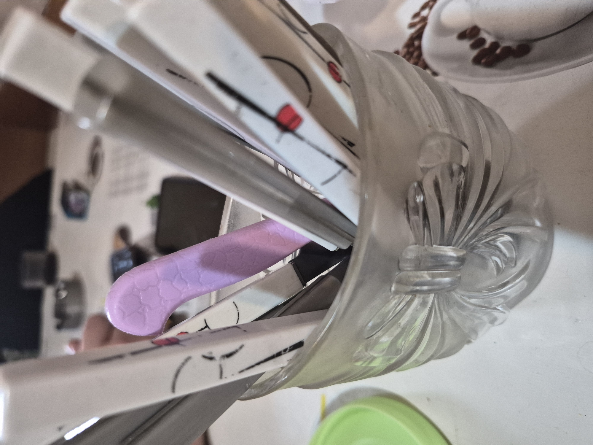 The image shows a close-up of a clear glass cup filled with various utensils and a singular light purple object with a textured surface. The cup has a decorative, sculpted design around its base. In the background, out of focus, are other objects on a white surface, including what appears to be an ashtray, a small dark electronic device, and some papers. There is also a blurred green object on the left side of the frame. The scene appears to be indoors, possibly on a table. The location in Yerevan, Armenia cannot be determined from the visual information provided.