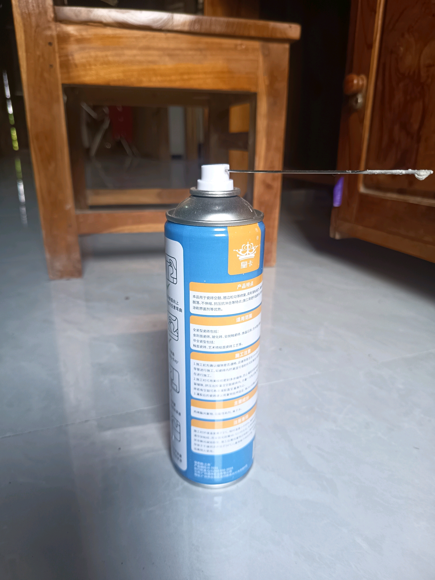 An eye-level, medium shot captures a close-up of a spray can with a long nozzle, which appears to be spraying a white substance. The spray can is blue and yellow and has Chinese writing on it. The can is positioned on a gray tiled floor. Behind the can, a wooden chair is visible, and to the right, a wooden cabinet with intricate carvings can be seen. The overall lighting is dim, with a slight sheen on the floor, suggesting indoor lighting. The location is not specified as Sidorejo, Indonesia.