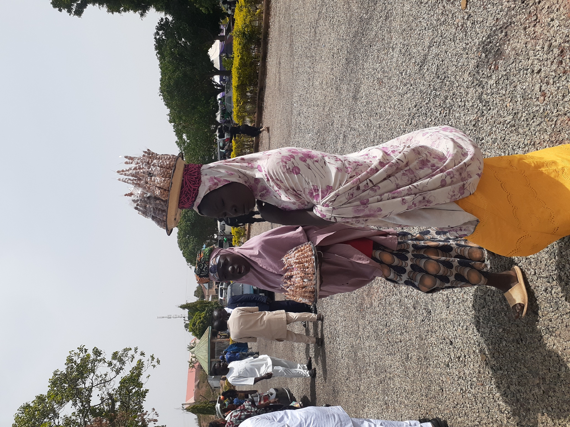 The image captures an outdoor scene featuring two women, likely vendors, selling packaged snacks or nuts in a bustling public area. The photograph is taken in bright daylight.
The first woman, positioned slightly behind and to the left, wears a light-colored garment with a floral pattern in shades of purple and pink, along with a matching head covering. She skillfully balances a wide, flat, light-brown tray on her head, which is piled high with numerous small, clear plastic packages. Each package appears to contain shelled nuts or similar snacks, arranged to form a conical mound. Her expression is calm as she looks towards the right. She also wears a plain yellow skirt or wrapper visible below her patterned top.
Standing in front of her and slightly to the right is a second woman, dressed in a light purple or pink head covering and a coordinating top. She holds a rectangular, flat, silver-colored tray at waist height, also filled with the identical individually packaged snacks. This woman is smiling warmly and looking directly at the camera, projecting a friendly demeanor. Her lower body is visible, showing dark patterned trousers with a distinctive light-colored circular design, and she wears simple, light-colored slip-on sandals.
The ground beneath them is a light-colored gravel or paved surface. In the background, several other people are visible, dressed in various casual and formal attire, suggesting a diverse crowd. Lush green trees and well-maintained shrubs line the background, with parked cars and the roofs of single-story buildings (some with green coverings) discernible further back under a clear, bright sky. The strong sun casts noticeable shadows, indicating it is likely midday or early afternoon. No visible text is present in the image.