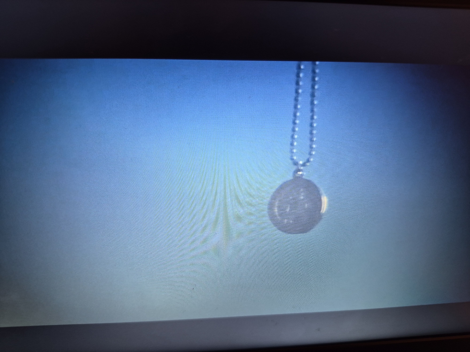The image shows a close-up view of a necklace with a circular pendant, hanging against a blurred blue background. The necklace is made of small, round beads. The pendant is a light-colored disk with a patterned surface. The background appears to be a gradient of blue, with lighter tones towards the bottom and darker tones towards the top, suggesting a soft light source. The image is framed by dark horizontal bands at the top and bottom, possibly indicating the edges of a screen or display. There are no people, actions, or interactions visible in the image. The scene is static and focused on the jewelry. The provided crops offer closer views of the necklace and pendant, as well as the textured background, highlighting the details of the beads and the pattern on the pendant. The location in the city of Sandwell, United Kingdom, cannot be determined from the visual information presented in the image.
