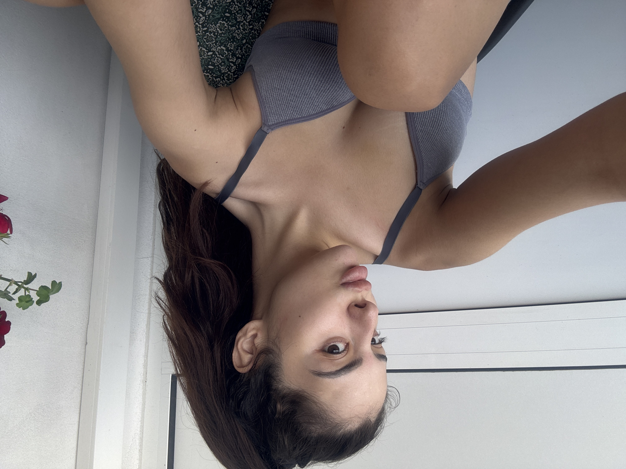 The image displays a young woman in an indoor setting, captured from an overhead perspective. The photo is rotated 90 degrees clockwise, so the woman appears to be lying on her back or side with her head facing upwards in the rotated view. She has medium-length dark brown hair and fair skin. Her eyes are open, and her lips are slightly parted, conveying a neutral expression. She is wearing a grey, ribbed sports bra or bralette.
Her upper body and arms are visible, with her arms bent and drawn towards her torso. Behind her upper back and head, a dark green fabric with a light floral pattern is visible, likely part of a pillow, blanket, or garment. In the bottom left corner of the frame (when correctly oriented), a partial view of a red flower with green leaves is present. The background consists of a bright white surface, possibly a wall or ceiling, with a distinct dark horizontal line, which could be part of a window frame or architectural molding. The lighting is bright, suggesting daytime conditions. The woman appears to be posing for the camera, possibly taking a selfie. There is no visible text in the image. The location context provided is Plovdiv, Bulgaria.