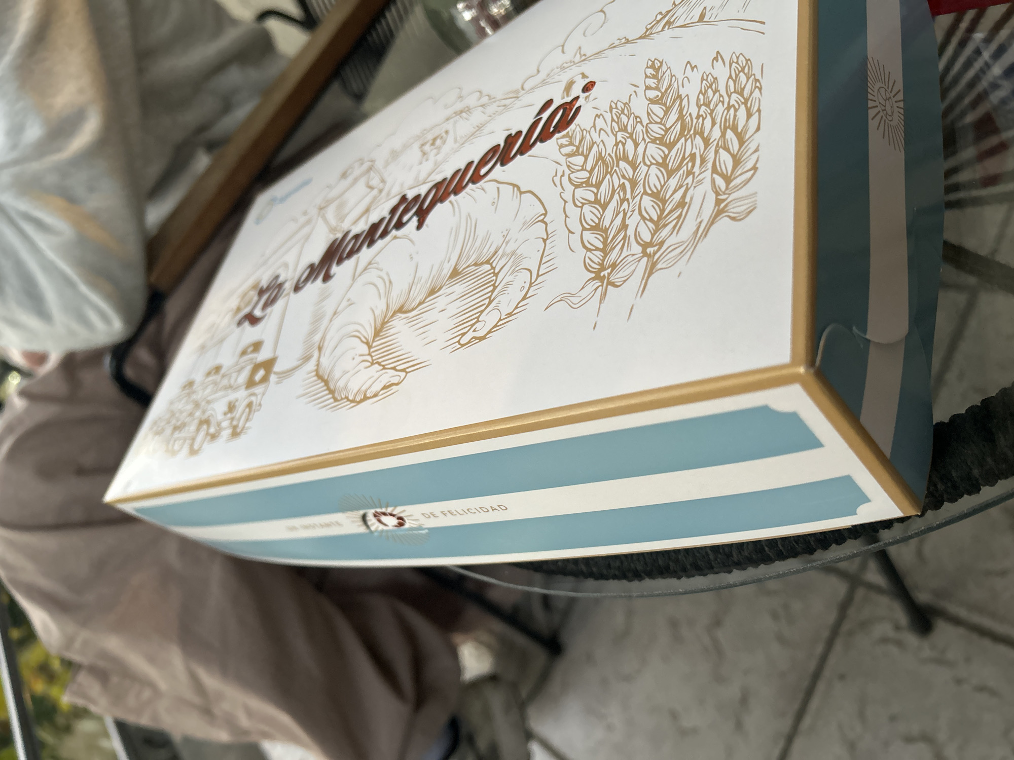 A rectangular white box with decorative printing is prominently displayed, tilted on a glass table. The box's top features a large, reddish-brown script text that reads "La Mantequería." Surrounding the text are intricate line art illustrations in a golden-brown hue, depicting a large croissant or pastry, stalks of wheat, and a faint cityscape or buildings in the background.
The sides of the box are adorned with light blue and white stripes, resembling the Argentinian flag, framed by a golden border. On one visible side, within the stripe pattern, a circular sun-like emblem is visible next to the text "DE FELICIDAD" (meaning "of happiness"). A partially visible word precedes "DE FELICIDAD," possibly "INSTANTE" or "FUENTE."
The background is softly blurred, showing a person wearing a light-colored top and beige or brown pants, suggesting an indoor or patio setting during daylight hours. The box likely contains baked goods or pastries from a brand named "La Mantequería," with packaging that incorporates elements of Argentinian national identity.
