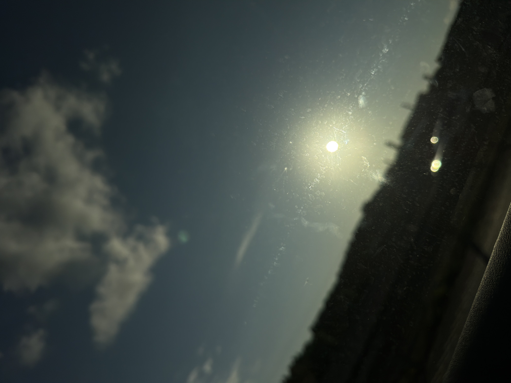 The image captures a sunlit sky with scattered clouds, observed through a dirty windshield. The sun is prominently featured, creating a bright glare. The lower portion of the image displays a blurred, dark landscape, possibly the side of a road or a hillside, with some faint structures visible in the distance. The overall impression is that of a view from inside a vehicle, looking outwards during daytime. There is no discernible location in Gothenburg, Sweden, or any specific activity or interaction depicted.