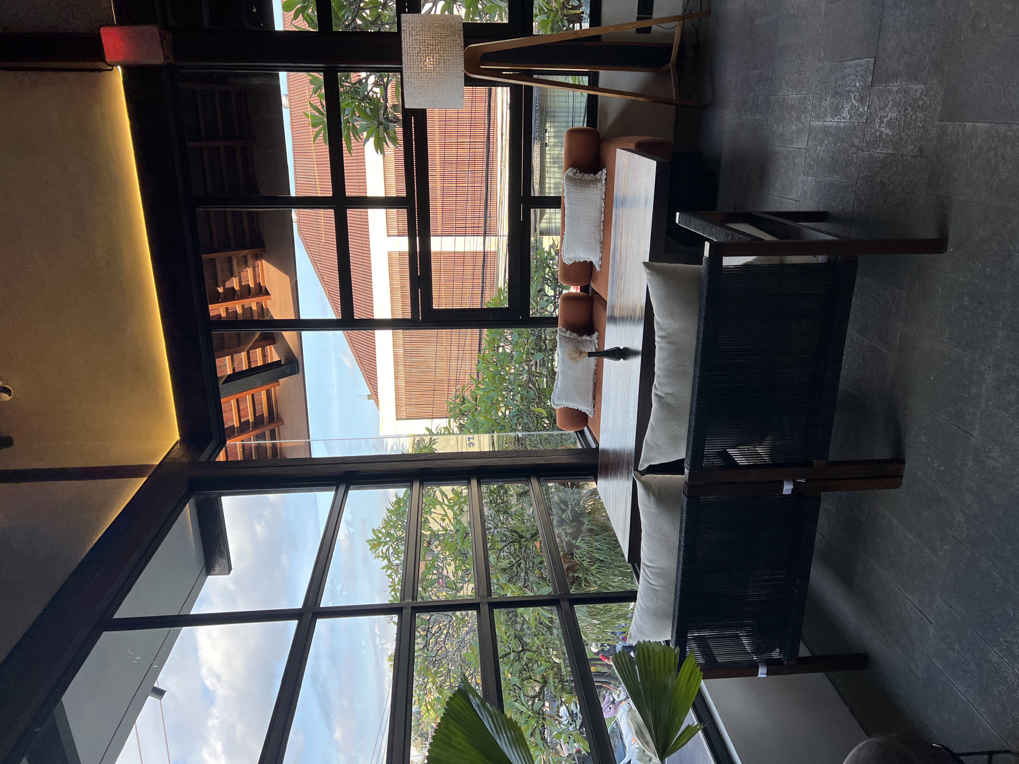 The image depicts an interior scene in Ubud District, Indonesia, featuring modern furniture, including a sofa and a chair, placed near a large window with a view of foliage and buildings. The scene is lit with a combination of natural light from the window and warm artificial light from a fixture. The interior suggests a café or a lounge area. The exterior view shows a mix of buildings, including ones with red tile roofs, and green trees, indicating a tropical setting. The floor is tiled in a dark gray or black color.